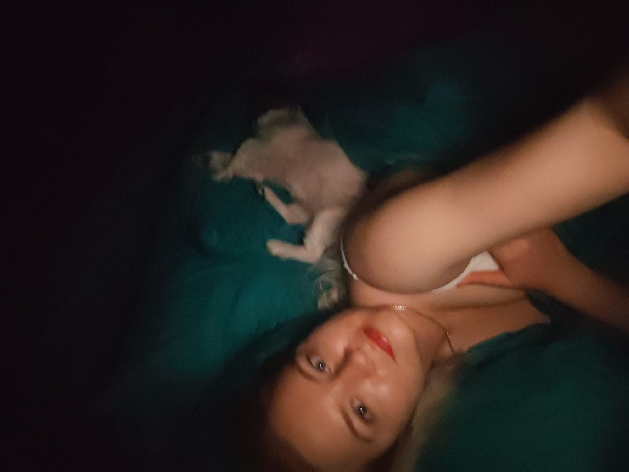 A woman with long blonde hair and a white top lies on a green bed next to a small white dog. The woman is wearing a gold necklace and red lipstick. The photo appears to be taken indoors, with dark lighting in the background. It is not possible to tell if this image was taken in the city of Izhevsk, Russia, from the given information.
