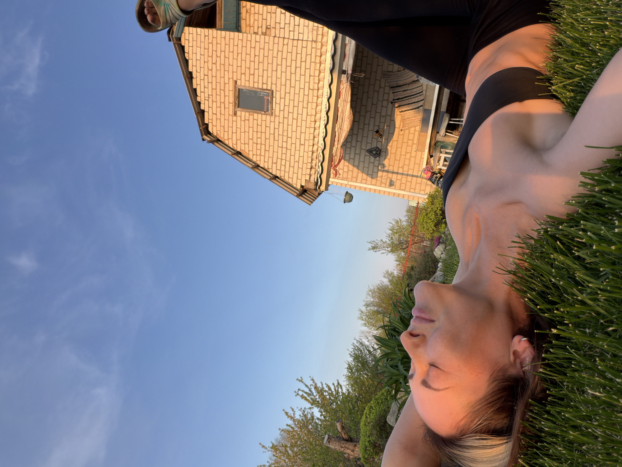 The image depicts a woman lying on green grass outside a two-story brick house under a blue sky. The woman has fair skin and a black top. She is lying on her back with her eyes closed, and her hands are behind her head. The house is a light tan color with a single window visible on the upper floor. There is a partially visible deck area next to the house. The sky is a clear blue with some wispy clouds. The image was taken at Kamyshin, Russia.