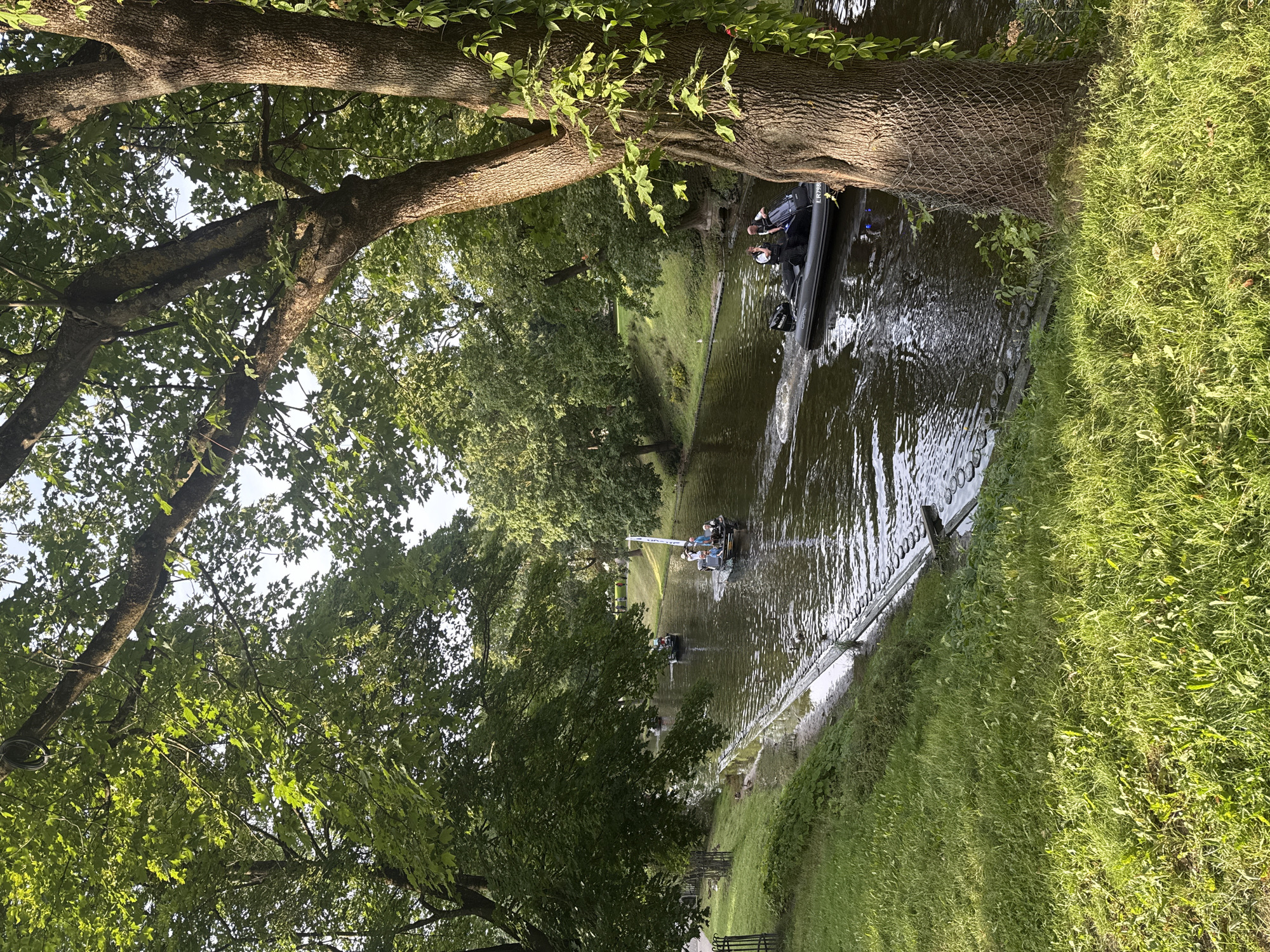 The image depicts a sunlit canal scene in Riga, Latvia, likely in the summer. Dense green foliage from mature trees overhangs a calm, dark body of water. The left bank is a grassy slope with a decorative metal fence at its base. The right bank features a large tree trunk covered partially by a wire mesh.
Several small boats navigate the canal. In the foreground, a dark inflatable boat with two individuals, one wearing a light-colored shirt and the other a dark shirt, is moving away from the viewer. The boat has an identification number "LR7998" visible on its stern. Further up the canal, another similar inflatable boat carries several people, some wearing light-colored shirts, and has a white flag on a pole. Another smaller boat is visible in the distance, further up the canal, with a few individuals on board. The water reflects the surrounding greenery and the sky. The overall atmosphere is peaceful and recreational.