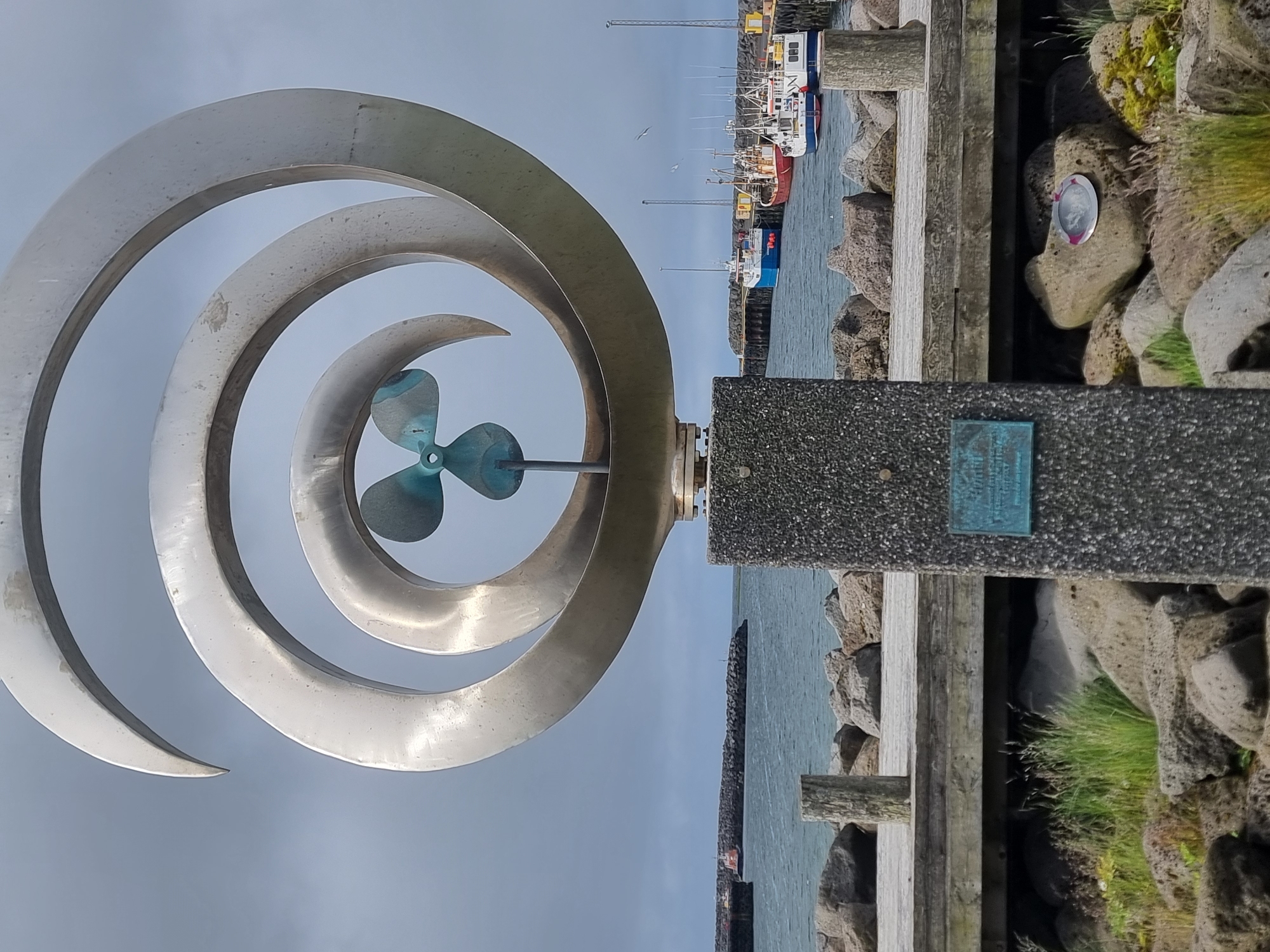 This is a photograph taken in Sandgerði, Iceland. The image features a prominent, large, spiraling metal sculpture with a three-bladed propeller at its center, mounted on a dark, speckled concrete pedestal. The sculpture is positioned on a waterfront with a wooden boardwalk in the foreground. Behind the sculpture, a body of water is visible, with several fishing boats docked in a harbor. The background shows a breakwater constructed of large rocks, and beyond that, a cloudy sky. The base of the sculpture's pedestal has a bronze plaque. Rocks and sparse vegetation are scattered around the base of the sculpture and the boardwalk. The overall scene depicts a coastal area with maritime elements and a significant public art installation.