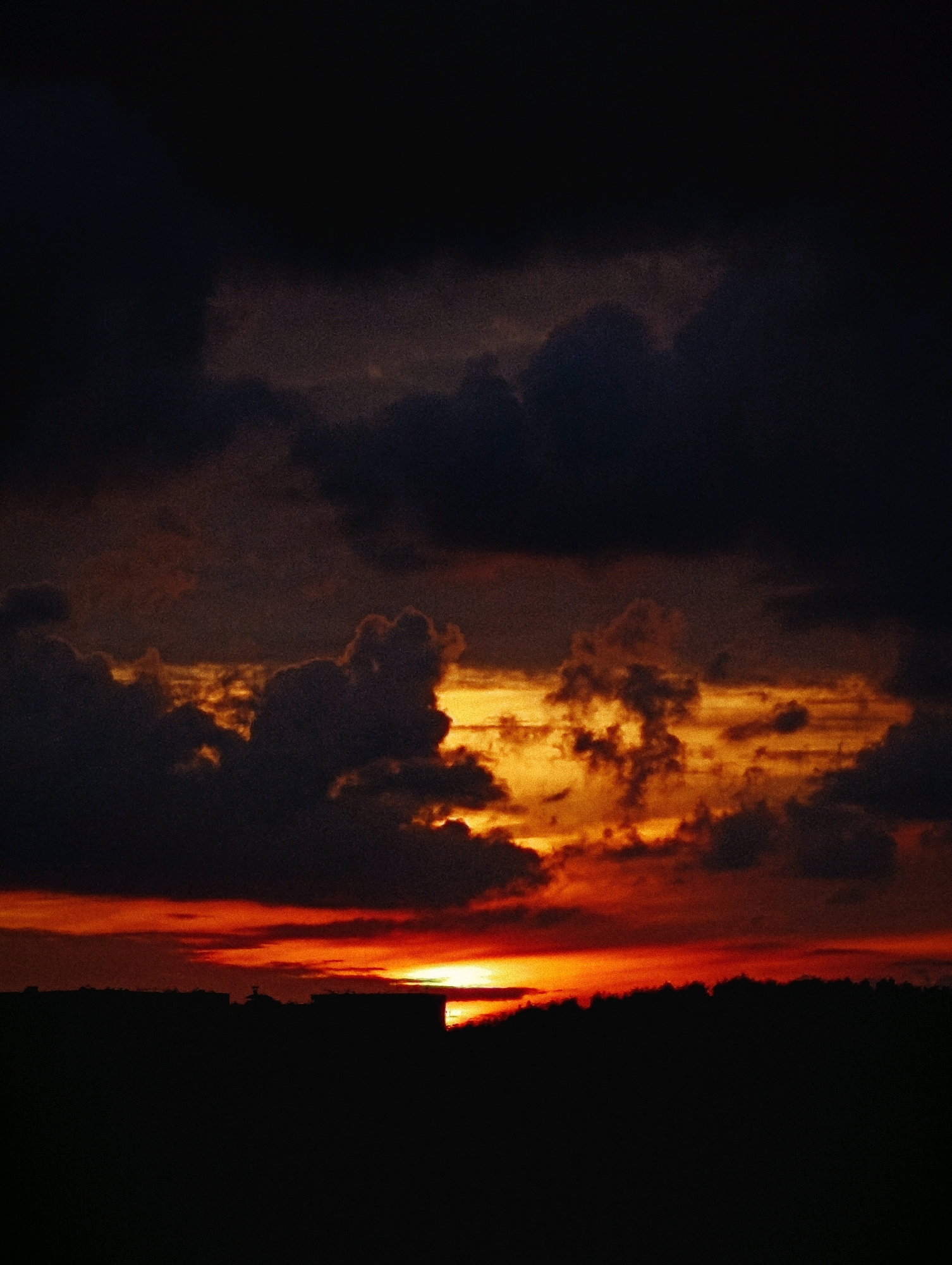 A dramatic sunset paints the sky in hues of orange and red, casting a fiery glow through dark, brooding clouds. The silhouette of a city skyline is visible along the bottom of the frame, its buildings rendered in deep shadow against the vibrant sky. The overall atmosphere is one of intense natural beauty and a touch of melancholy.