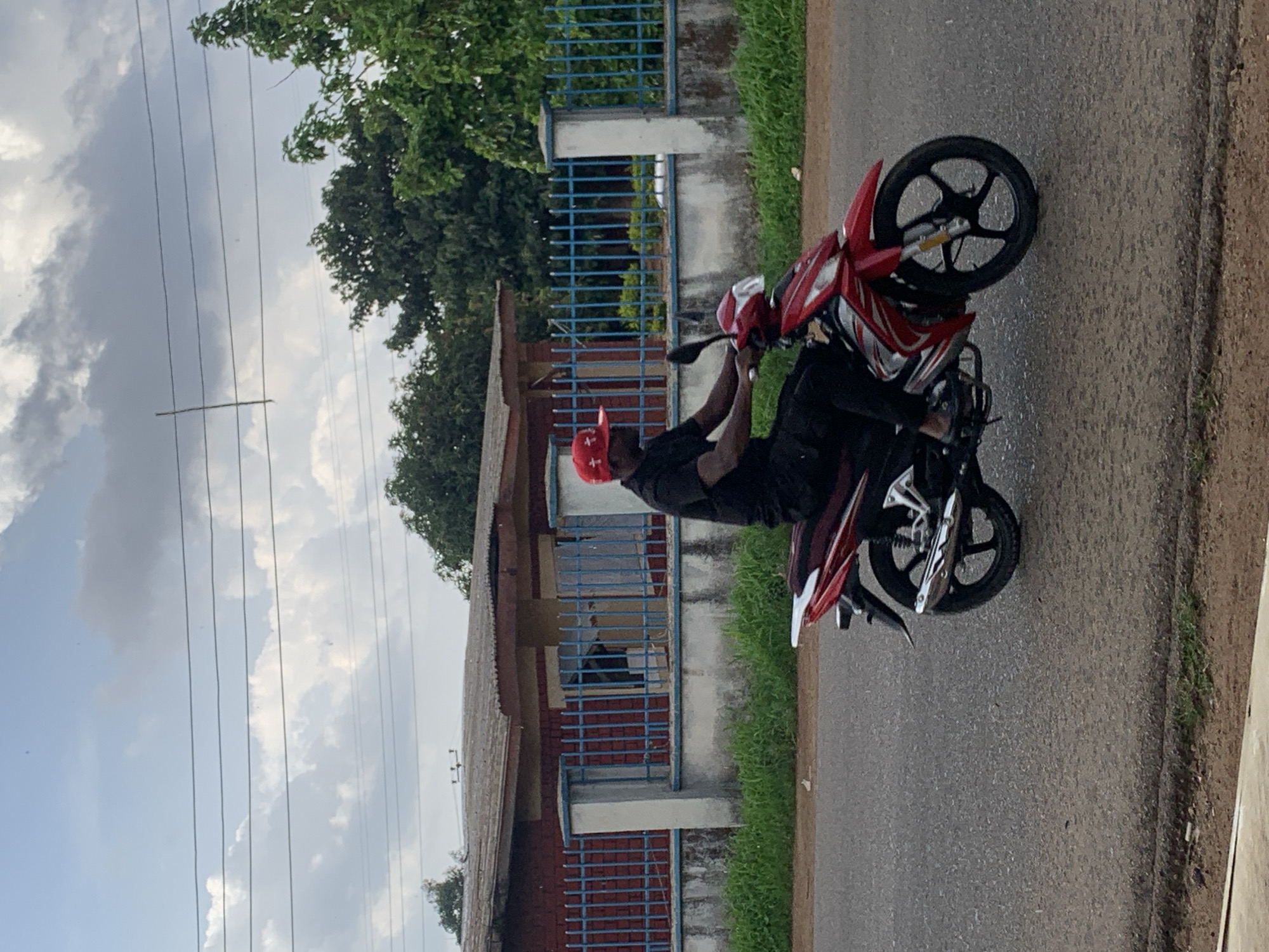 A vertical outdoor shot captures a person on a red and black motorcycle, appearing to perform a slight wheelie or holding the front wheel elevated. The individual is wearing a red cap and dark clothing, seated on the motorcycle with hands on the handlebars. The motorcycle features black wheels with silver spokes and red fairings.
The scene takes place on an asphalt road. In the background to the left, there's a building with a corrugated roof, reddish-brown walls, and light blue metal grates over windows and fences. Green grass grows along the road and near the building. Densely foliated green trees are visible in the middle and right background. The sky is partly cloudy, with a mix of grey clouds and lighter patches, traversed by several parallel power lines. The overall lighting suggests it is daytime, under overcast or partly cloudy conditions. No discernible text is visible in the image. The setting appears to be a typical roadside in a residential or semi-urban area.