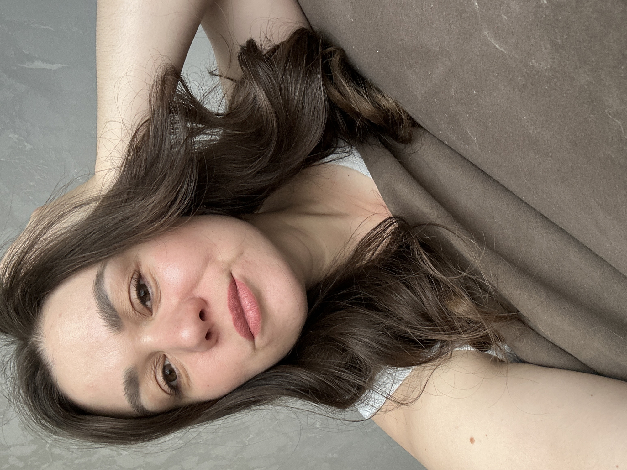 A young woman is captured in a close-up, horizontally oriented portrait, appearing to be lying down. She looks directly at the camera with a soft, neutral expression, possibly a faint smile, her head tilted slightly. She has long, wavy dark brown hair that cascades around her face and over her shoulders and visible arm. Her eyes are brown, and her eyebrows are dark and well-defined. She is wearing light pink lipstick. Her fair skin appears smooth, and a small mole is visible on her upper arm. A strap of a light-colored or white top is visible near her shoulder. The background features a textured grey surface on the left, possibly a wall or floor, and a dark brown fabric, resembling a blanket or upholstery, on the right. The brown fabric shows some small specks or lint. The lighting is soft and even, indicating an indoor setting. No text is visible in the image.