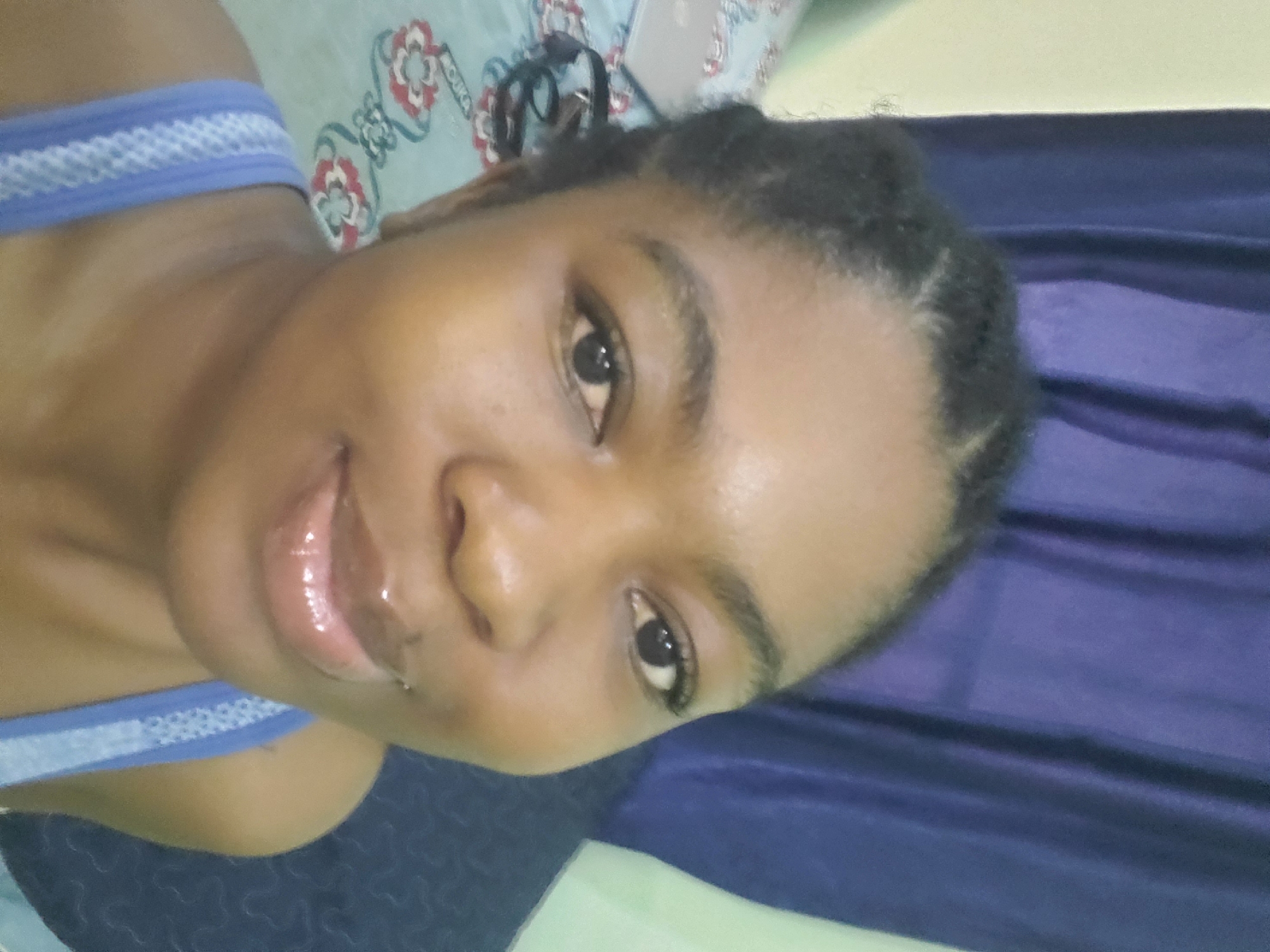 The image is a close-up selfie of a dark-skinned young woman with dark hair styled in small braids. She is wearing a blue and white patterned top. Her lips appear glossy. The background includes a blue draped fabric, a light green surface, and a patterned pillow. The location in the city of Barden Yana, Nigeria cannot be determined from the image.