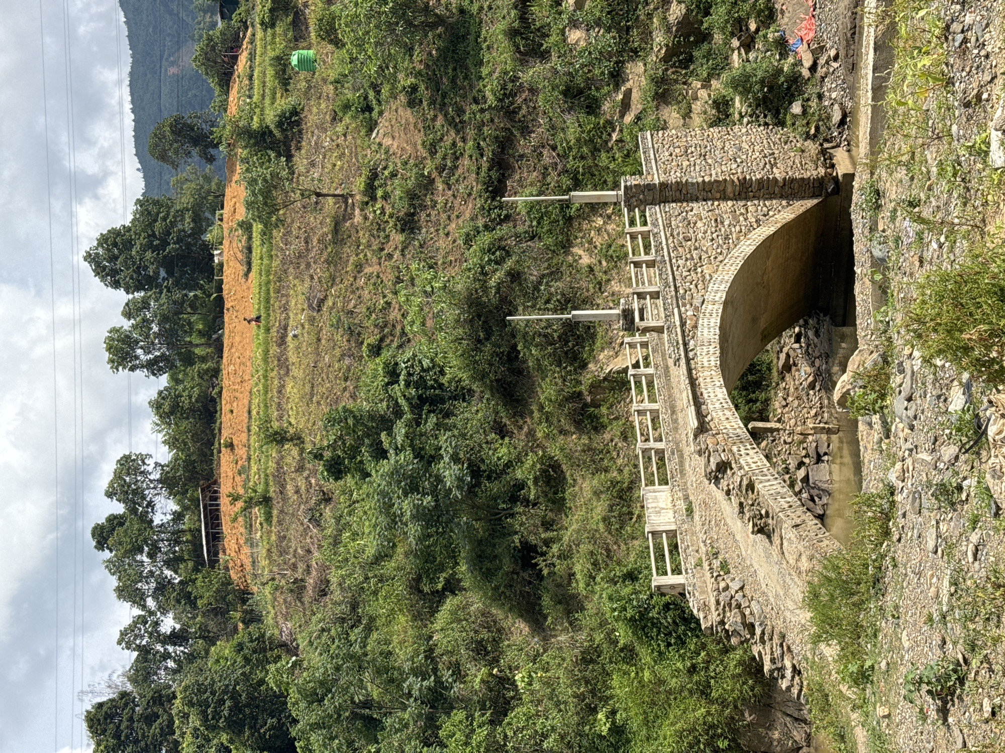 This image presents a serene, mountainous landscape, likely situated in a rural area like Khao Mang, Vietnam. In the foreground, a robust stone arch bridge spans a narrow, shallow stream. The bridge features stone walls built with irregular rocks and includes concrete railings with lighter-colored, possibly wooden, posts forming a balustrade along its length.
To the left of the bridge, a steep hillside dominates the scene. It is a mix of lush green trees and dense foliage, interspersed with patches of cultivated land featuring light brown, tilled soil. A small, distant figure, possibly a person, can be seen on one of these cultivated sections. Further up the hill, partially obscured by trees, is a simple shack or shelter with a light-colored roof. A bright green plastic container, perhaps a water tank, is visible higher on the right side of this slope.
The background is characterized by rolling mountains under a bright, overcast or partly cloudy sky. Utility lines stretch across the upper left portion of the frame. The overall lighting suggests it is daytime, possibly morning or afternoon, with soft, diffused light. The scene depicts a harmonious blend of human infrastructure and agricultural activity within a natural, rugged mountain environment, devoid of any visible text.