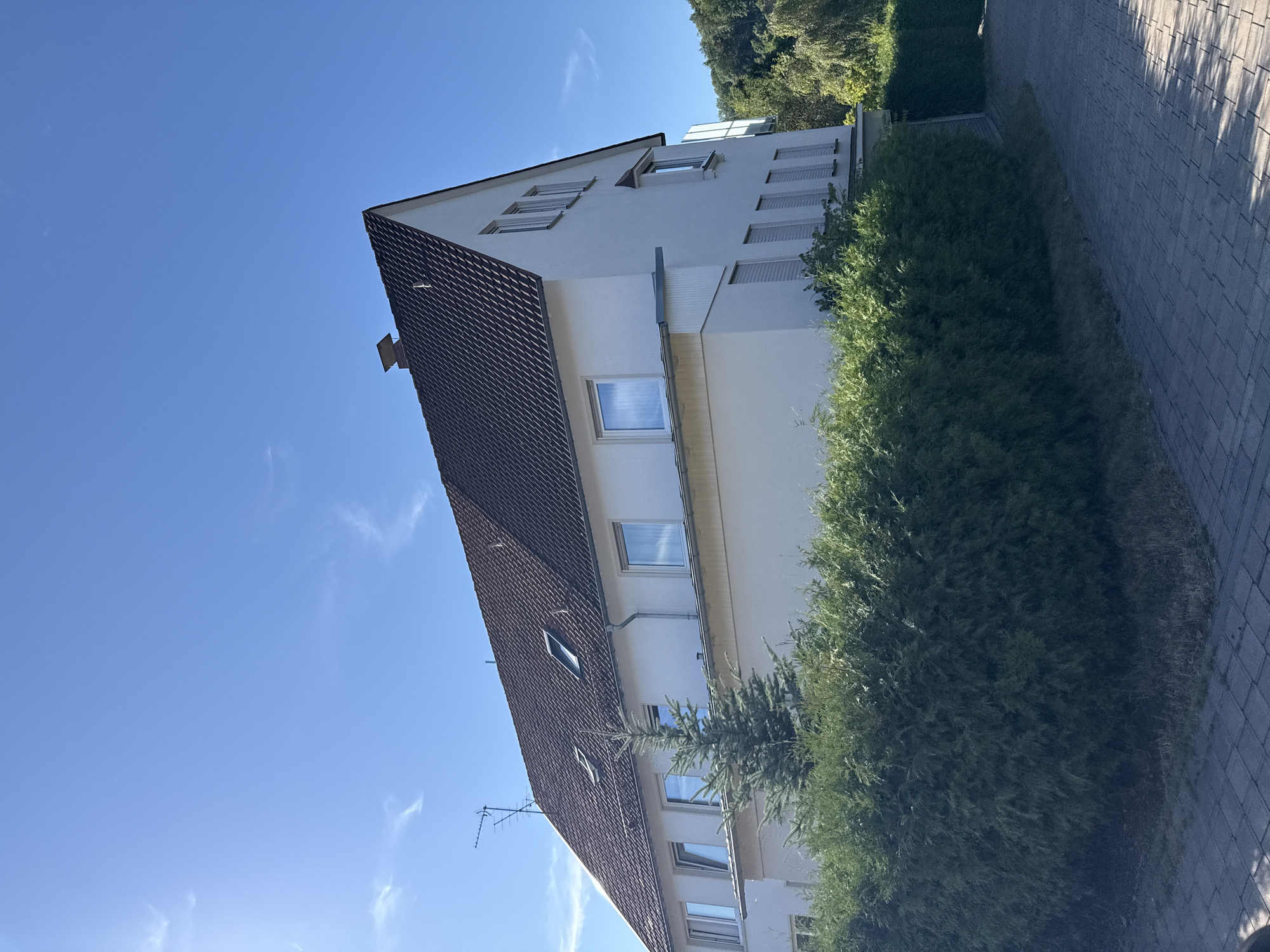 The image shows a white, multi-story building with a dark tiled roof, located in the city of Walkenried, Germany. A large, dense green hedge runs along the front of the property, partially obscuring the lower level of the building. A paved walkway is visible in the foreground. The sky is clear and blue, with a few wispy clouds. A small evergreen tree stands in front of the hedge. The building has several windows, some with reflections of the sky and trees. An antenna is mounted on the roof. The overall impression is a residential setting on a sunny day.