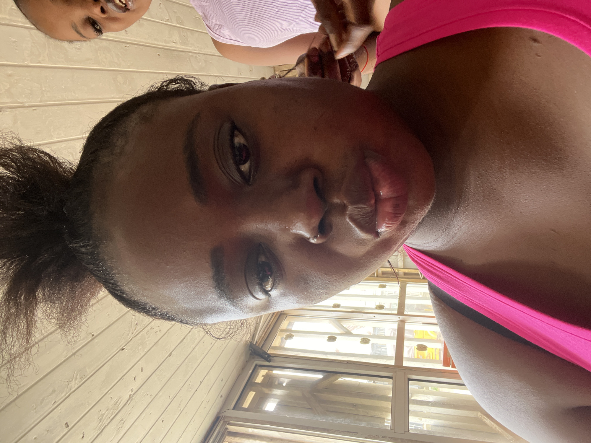 A close-up, selfie-style shot features a dark-skinned woman in the foreground, facing the camera with her mouth slightly pursed. She has her dark hair pulled up in a messy bun and is wearing a bright pink tank top. Her skin appears slightly shiny, possibly from sweat. In the blurred background, a second dark-skinned woman, wearing a light purple, ribbed tank top, is partially visible, looking towards the left. The setting appears to be indoors with wooden paneling on the ceiling and a window with vertical blinds in the background. The overall lighting is bright and natural. The image is not taken in Abuja, Nigeria.
