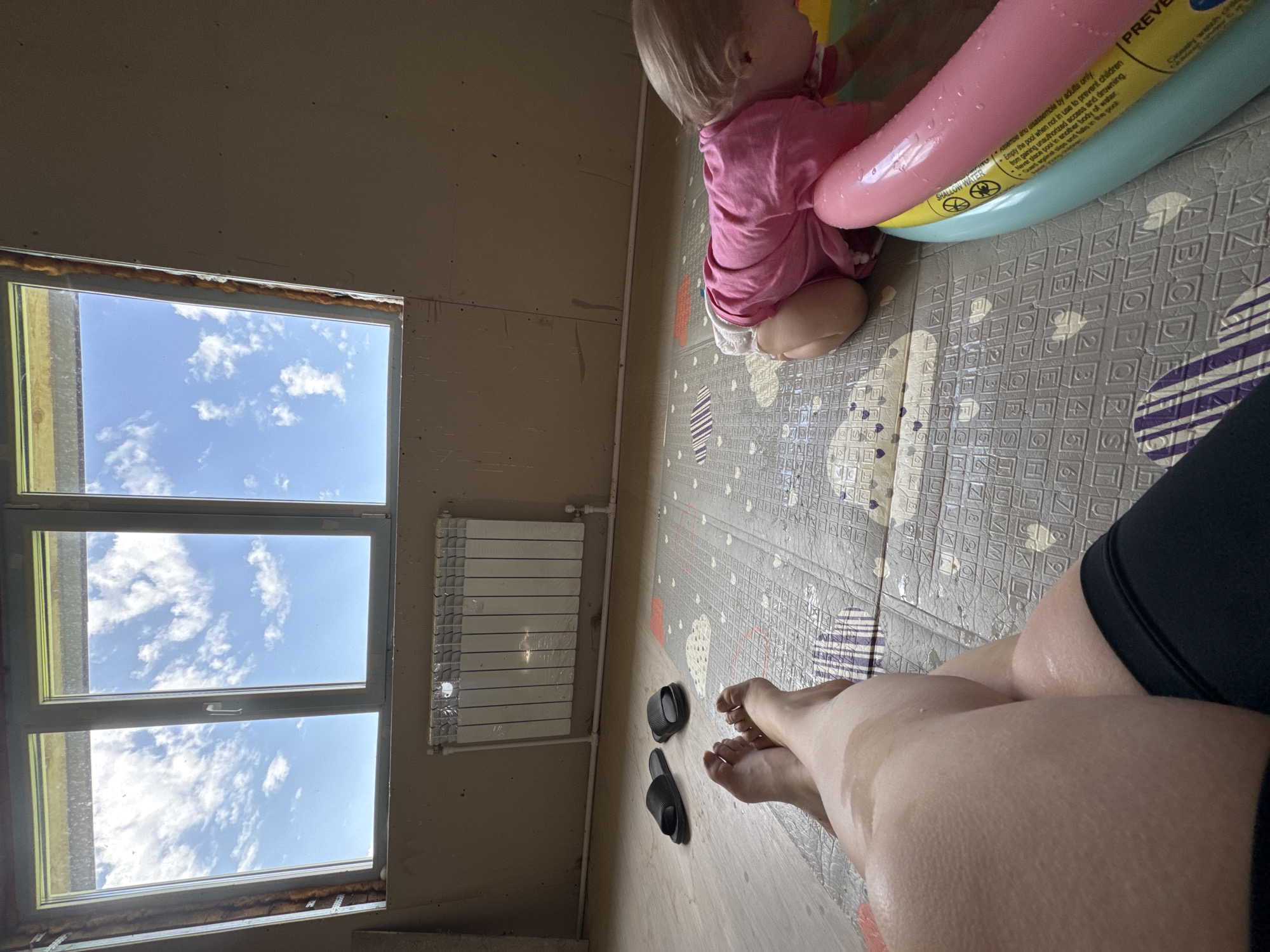 The image shows an interior scene with a baby in a small inflatable pool, a partially visible person, a window overlooking a blue sky with clouds, and a radiator. The location is Фоки, Russia.
* **People:** A baby wearing a pink shirt is sitting in an inflatable pool. A person's legs and feet are in the foreground, suggesting the image is taken from their perspective.
* **Subjects:** The baby, the person, the inflatable pool, the window view.
* **Objects:** Inflatable pool, window, radiator, sandals, floor mat with a pattern.
* **Actions:** The baby is sitting in the pool. The person is observing the scene.
* **Interactions:** The person is likely supervising the baby in the pool.
* **Environment:** The room appears to be under construction, with unfinished walls. The window offers a view of a bright, cloudy sky.