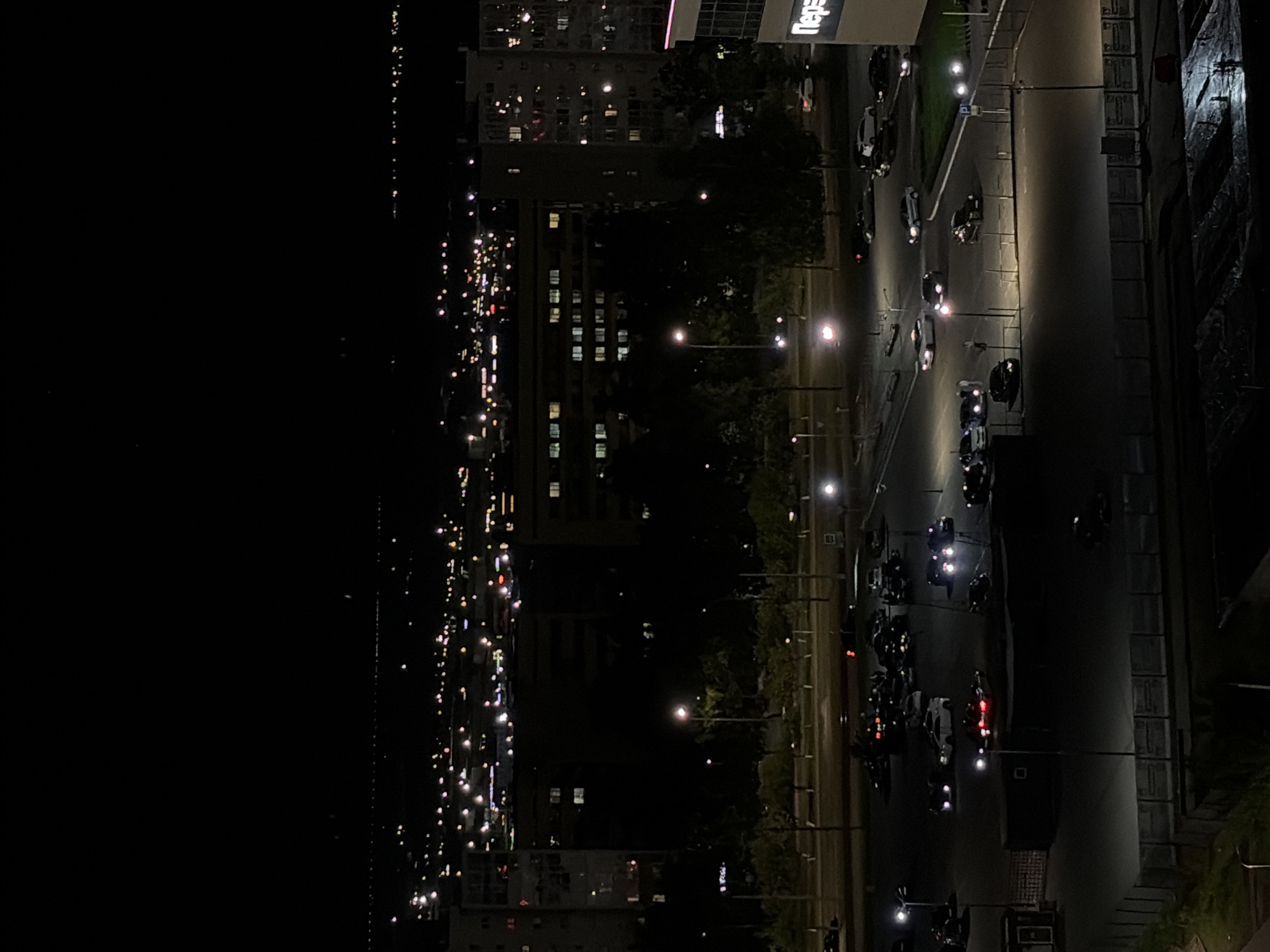 An evening view from an elevated position overlooking Naberezhnye Chelny, Russia. A roadway runs near the vantage point, with cars parked and moving. Further away, city lights indicate buildings and streets. In the far distance, a body of water reflects light.