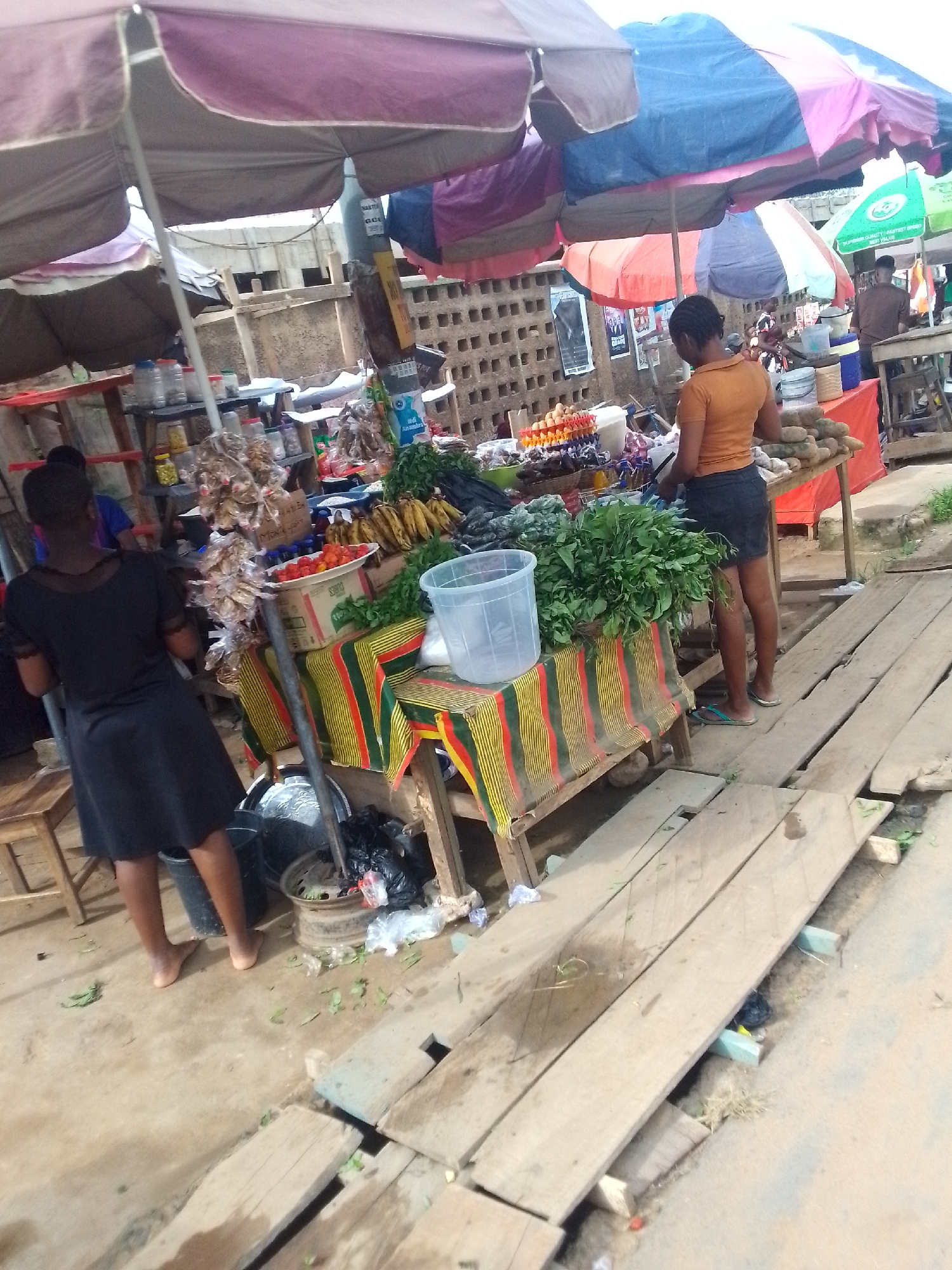 The image depicts a bustling open-air market scene in Egbagu, Nigeria. Two women are visible: one dressed in a black dress, barefoot, and the other in an orange top and dark shorts, wearing flip-flops. They stand near tables laden with produce including bananas, tomatoes, yams, and leafy green vegetables. Merchandise such as spices and dried goods are also displayed. The tables are covered with colorful striped cloths. The market stalls are sheltered by a series of large, colorful umbrellas. The ground is uneven, with wooden planks laid down as a makeshift walkway over dirt or concrete. Litter and debris are scattered around the immediate vicinity. In the background, a concrete block wall is visible.