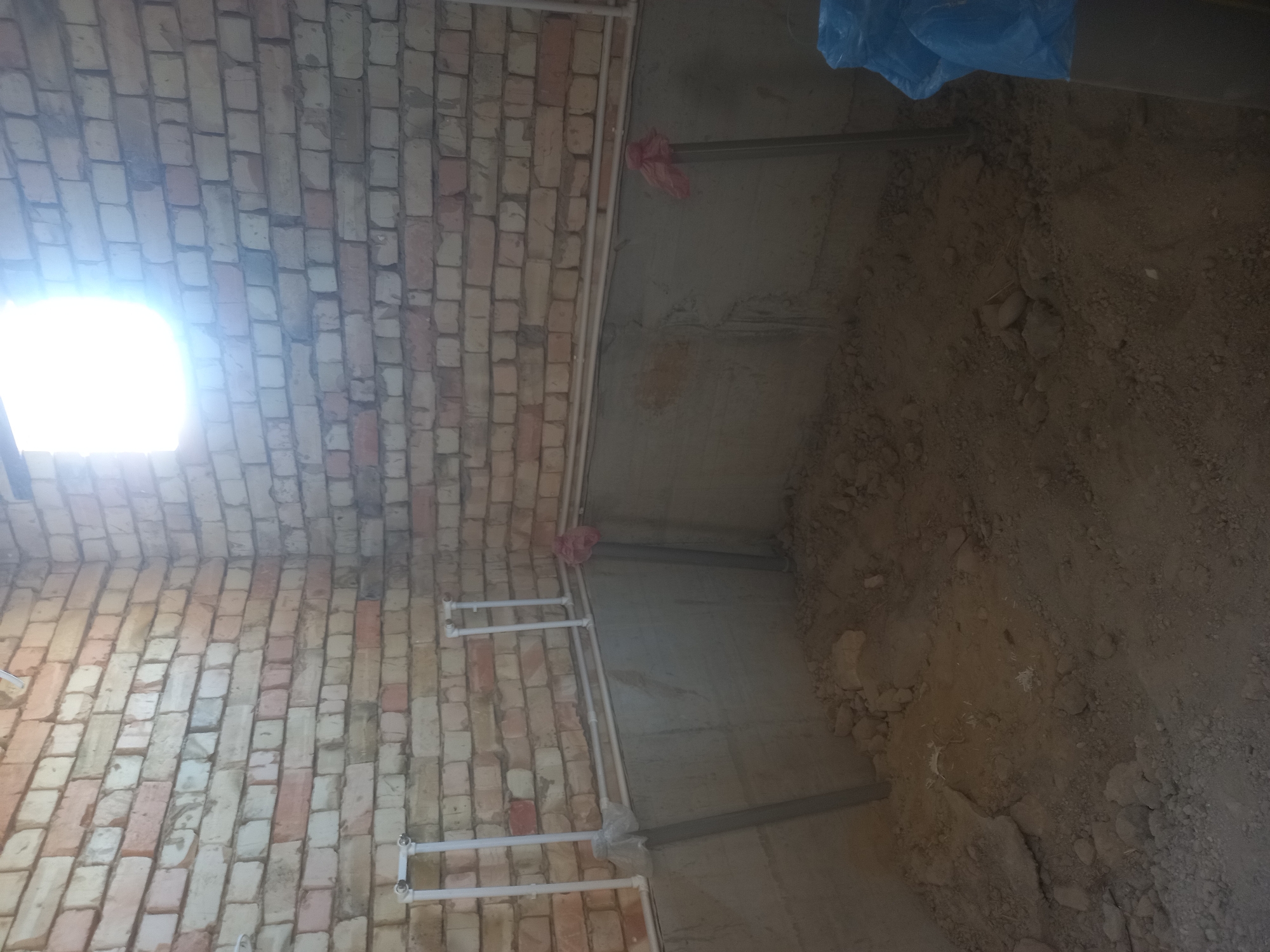 The image depicts an unfinished interior space, likely a room under construction, with exposed brick walls and a concrete foundation. In the background, a bright light shines from a window or opening in the upper right portion of the frame. White PVC pipes are visible along the walls, suggesting plumbing installation. The floor is covered with dirt and debris, indicating that work is ongoing. In the foreground, a blue plastic bag is partially visible on the right side. The scene is set in Bulungur, Uzbekistan, a city known for its agricultural and industrial activities.