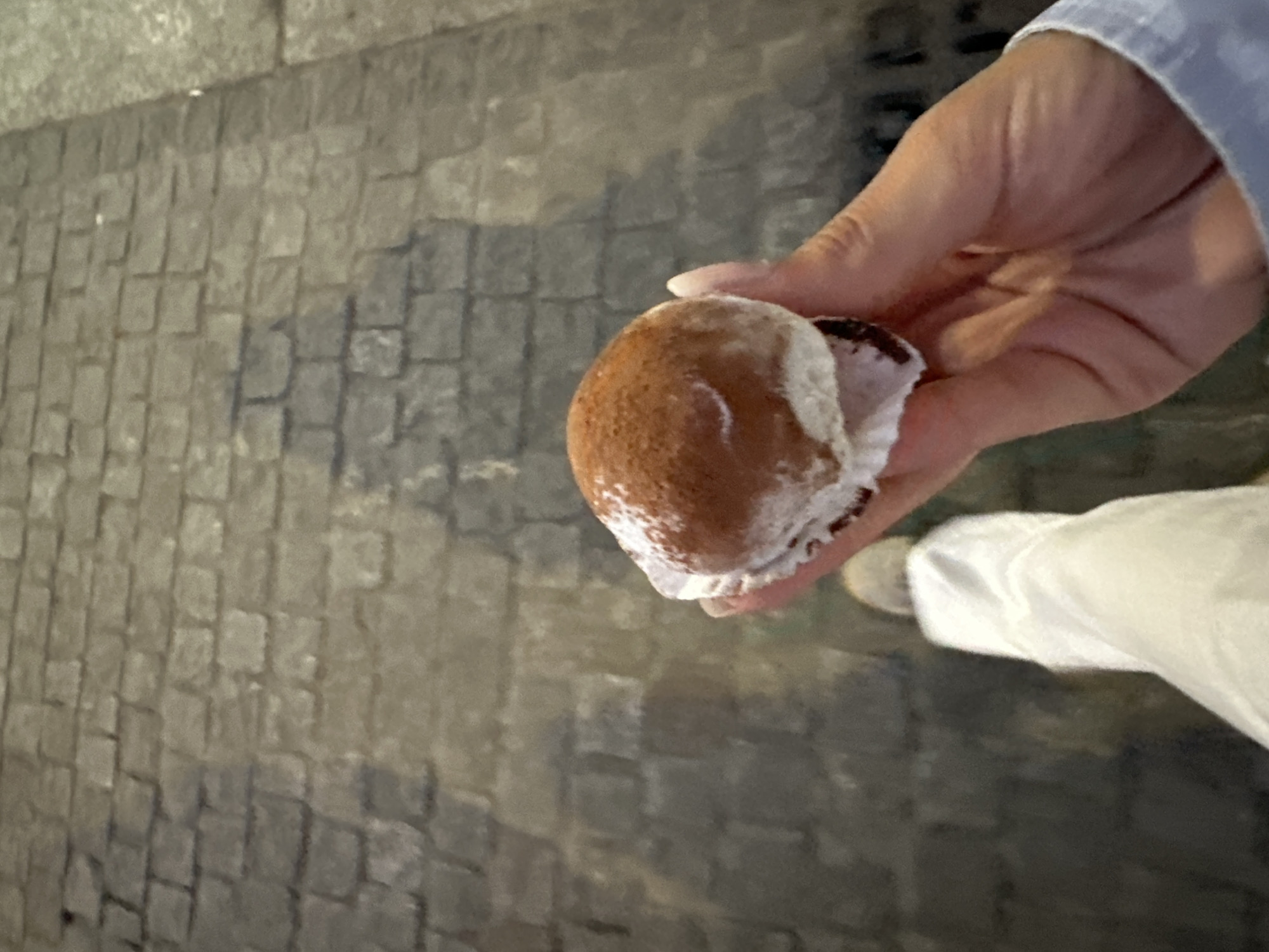 A hand holds a Maritozzo pastry in front of a cobblestone street. The pastry is dusted with powdered sugar. Part of a white garment is visible on the lower right. The location is likely Milan, Italy, as Maritozzo pastries are a typical Roman pastry.