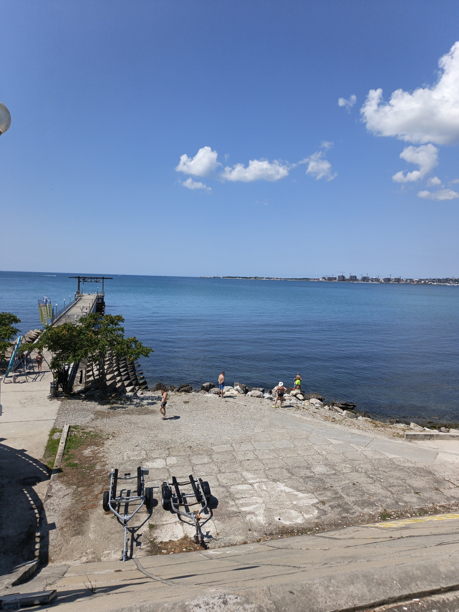 In the city of Pshada, Russia, on a bright sunny day, a wide shot captures a coastal scene. The clear blue sky is dotted with a few fluffy white clouds. A long, wooden pier extends out into the calm, azure sea. On the shore, a pebbled area transitions to a concrete ramp. Two boat trailers are parked on the concrete. Several people are scattered across the beach and along the water's edge. Two individuals are standing near the water, and a few others are visible further down the pier. The background features a distant shoreline with buildings and construction cranes. The overall ambiance is one of a peaceful summer day at the seaside.