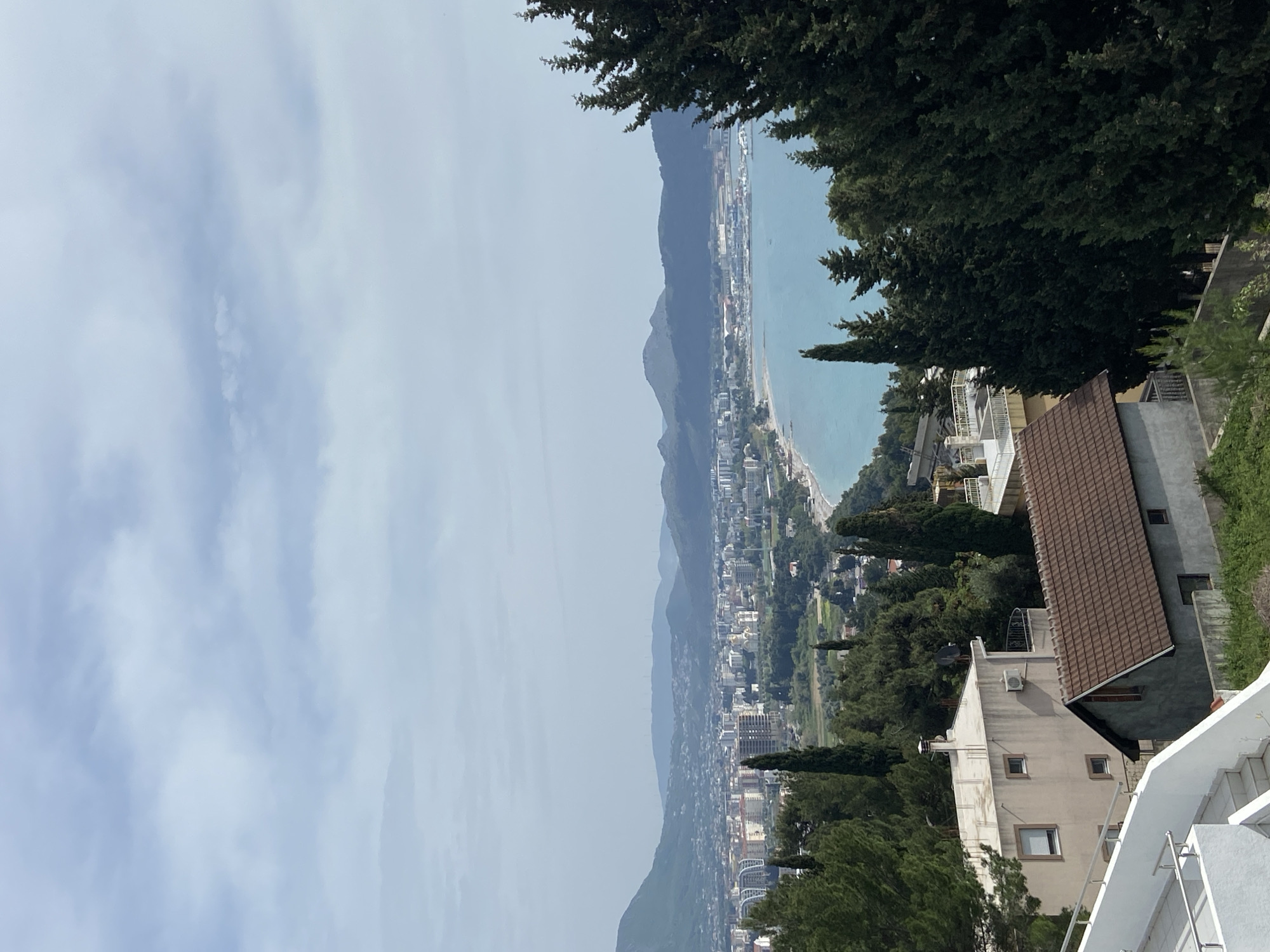 The image presents an expansive elevated view of a coastal landscape, likely captured on an overcast or hazy day. The primary focus is a bustling coastal town situated along a bay, flanked by mountains.
In the foreground, on the right side, residential buildings with a mix of light-colored walls and tiled roofs are visible, nestled among numerous tall, dark green trees, possibly cypress. A white staircase with a metallic handrail ascends on the far right, suggesting a terraced or sloped terrain.
The midground features a long stretch of urban development forming the coastal town of Šušanj, Montenegro. Numerous buildings of various sizes and architectural styles densely populate the area between the sea and the foothills of the mountains. Green vegetation is interspersed throughout the town, adding to the landscape.
To the left of the town, a tranquil, light blue body of water, presumably the Adriatic Sea or a bay, extends towards the horizon. A distinct coastline and narrow beach area are discernible where the town meets the water. Beyond the water, a prominent range of dark mountains rises steeply, running parallel to the coast and disappearing into the atmospheric haze.
The sky occupies the upper portion of the scene, appearing largely overcast with diffused light, indicating a daytime setting. No individuals or specific activities are observable, and no visible text is present. The overall impression is a scenic panoramic overlook of a coastal resort town.