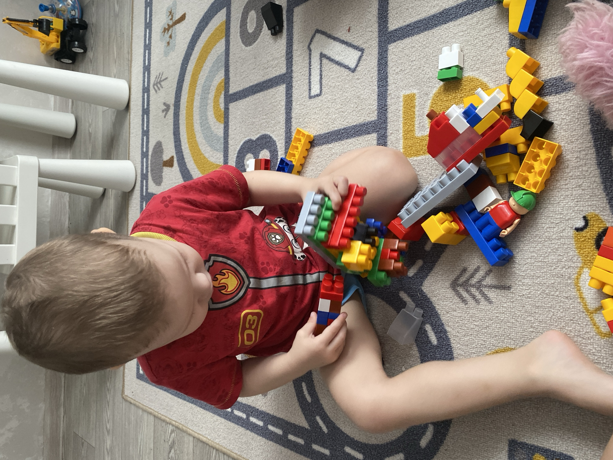 A single young individual, presumed to be a child, is observed indoors, seated on a light-colored rug with a decorative pattern featuring a rainbow, road lines, trees, and numbers (e.g., '7', '8', '9'). The individual wears a red short-sleeved t-shirt with a 'Paw Patrol' emblem and the number '03' on the chest, along with blue shorts. The individual's gaze is directed downwards towards a collection of multi-colored plastic building blocks, several of which are being manipulated by both hands.
Numerous building blocks in red, blue, yellow, green, white, gray, black, and brown are scattered across the rug, some interconnected to form structures, and others loose. A small toy figurine, appearing to be a person in a green cap, is visible among the blocks. In the upper right background, a small yellow toy excavator truck and a blue spinning top are present on a light-colored wooden floor. White, cylindrical furniture legs, likely from a table or chairs, are also visible in the background. A light-colored wall forms the farthest background element. A small, clear plastic container and a pink fluffy object are visible in the lower right area of the frame. The scene is illuminated by ambient indoor light. This scene is located in Sochi, Russia.