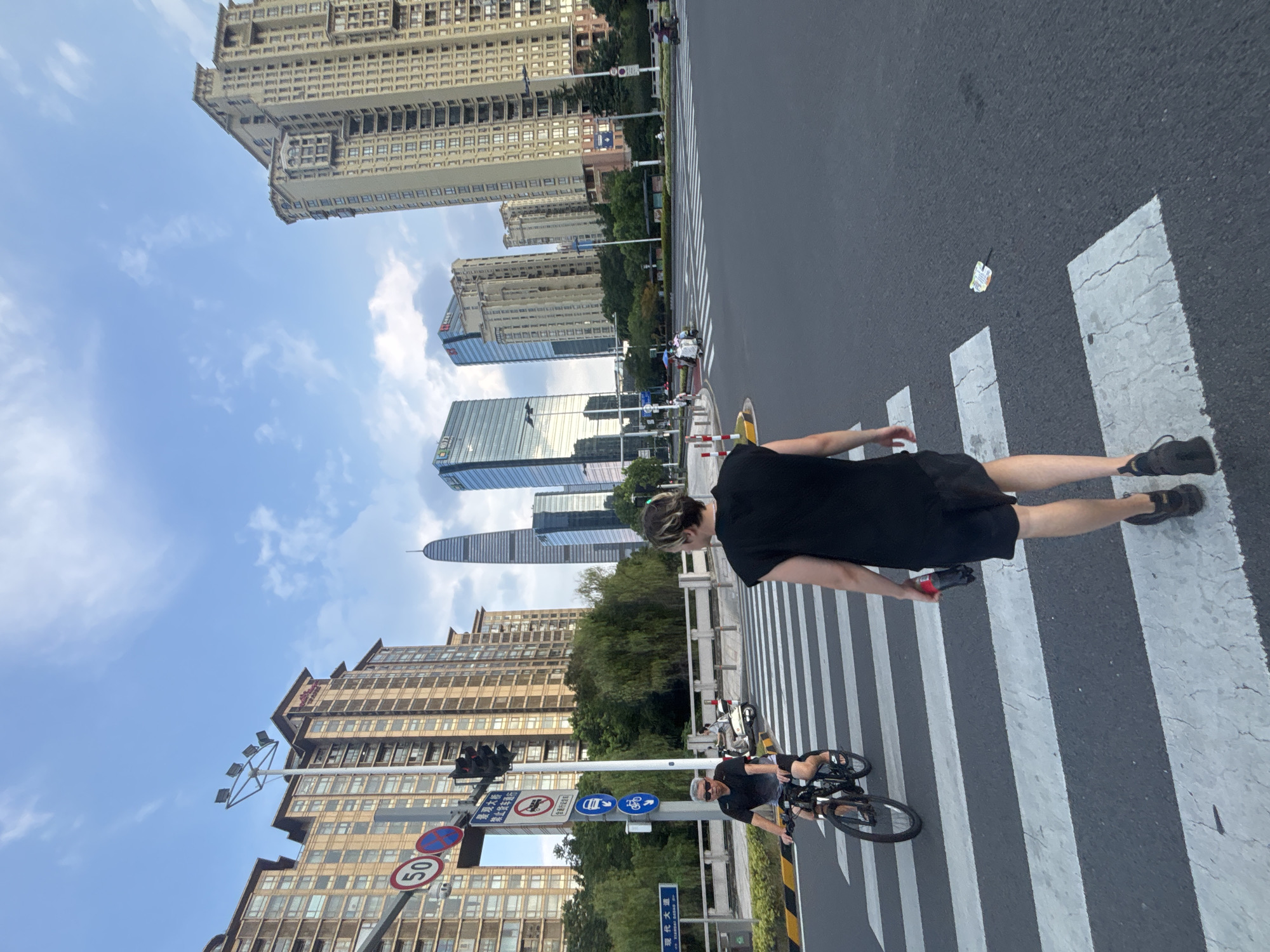 The image depicts a street scene in Suzhou Industrial Park, China. A pedestrian, wearing a black tank top and shorts, walks across a crosswalk carrying a red bottle in their right hand. To the left, a man on a bicycle, wearing a black t-shirt and shorts, rides on the road. The crosswalk is marked with white stripes on a dark asphalt road. In the background, tall buildings with many windows line the street, and trees are visible between the buildings and the road. Traffic lights and road signs are also present, including a speed limit sign showing "50". The sky is blue with scattered clouds. The overall impression is an urban environment during daylight.