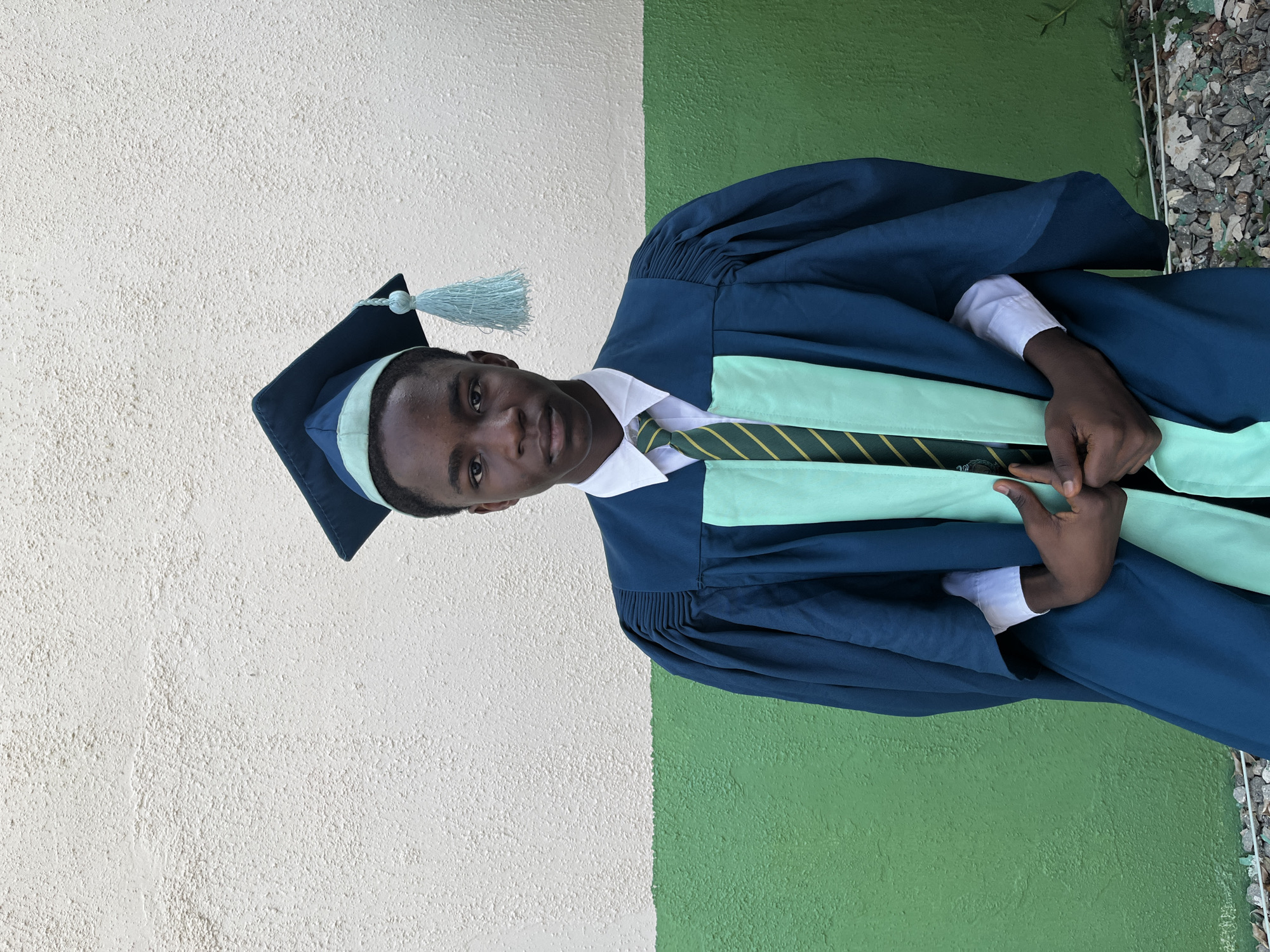A young, dark-skinned man stands in a graduation gown and cap with a light green sash and tie, posing for a photo. He is wearing a white collared shirt underneath the gown. The gown and cap are dark blue. He is in front of a wall that is half white and half green, possibly on a campus in Jos, Nigeria. The location is an outdoor setting with some scattered gravel visible near the bottom of the frame.