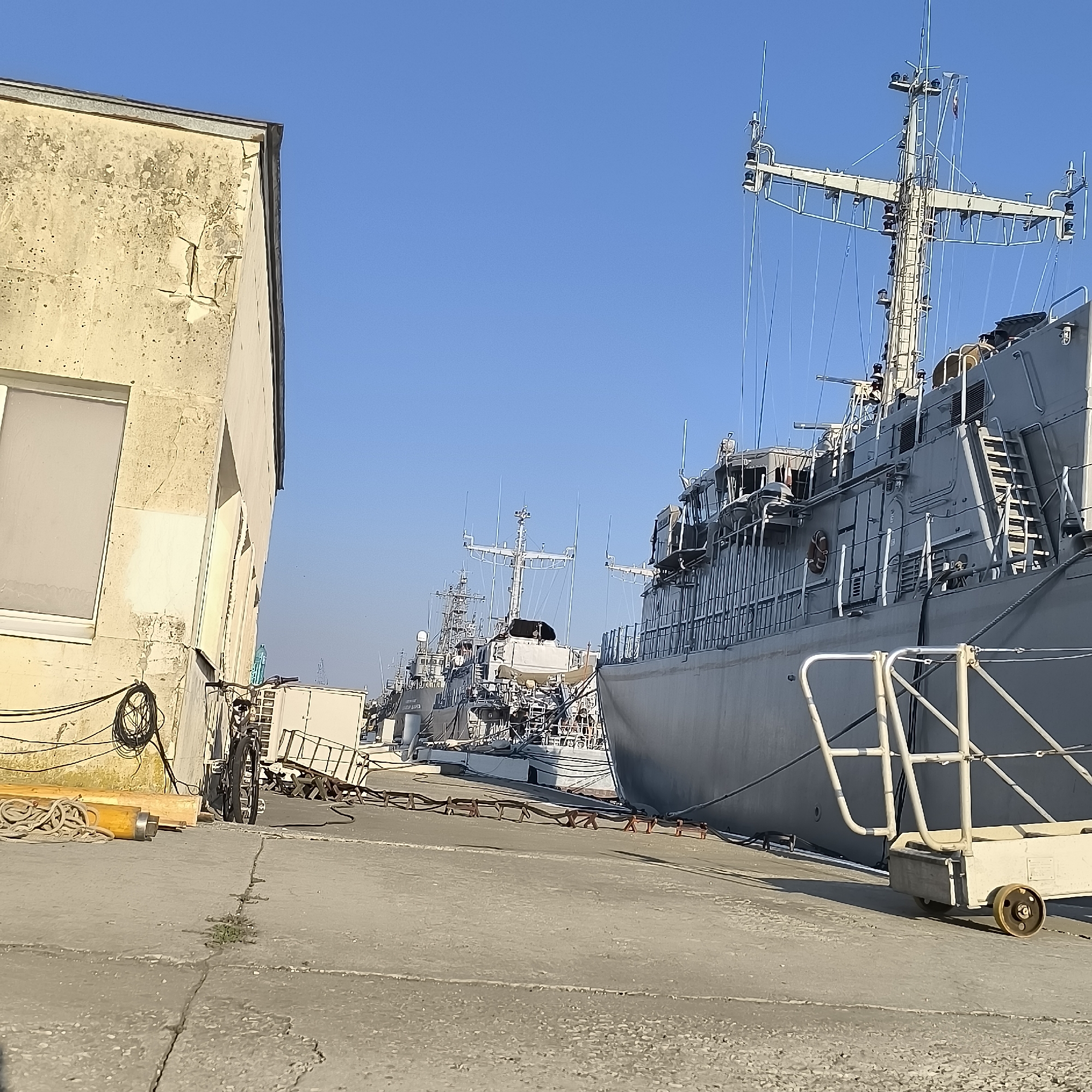A port scene in Varna, Bulgaria, features several gray naval vessels docked along a concrete pier under a clear blue sky. In the foreground, a weathered, light-colored building with a large, opaque window stands to the left. A black bicycle is parked near the building. Ropes and a yellow cylindrical object are placed on the ground. The primary focus is on the imposing hull of a large gray ship to the right, with its mast and rigging visible against the sky. Several other similar ships are lined up behind it. A white cart with wheels is positioned on the pier in front of the large ship. The environment is bright and sunny.