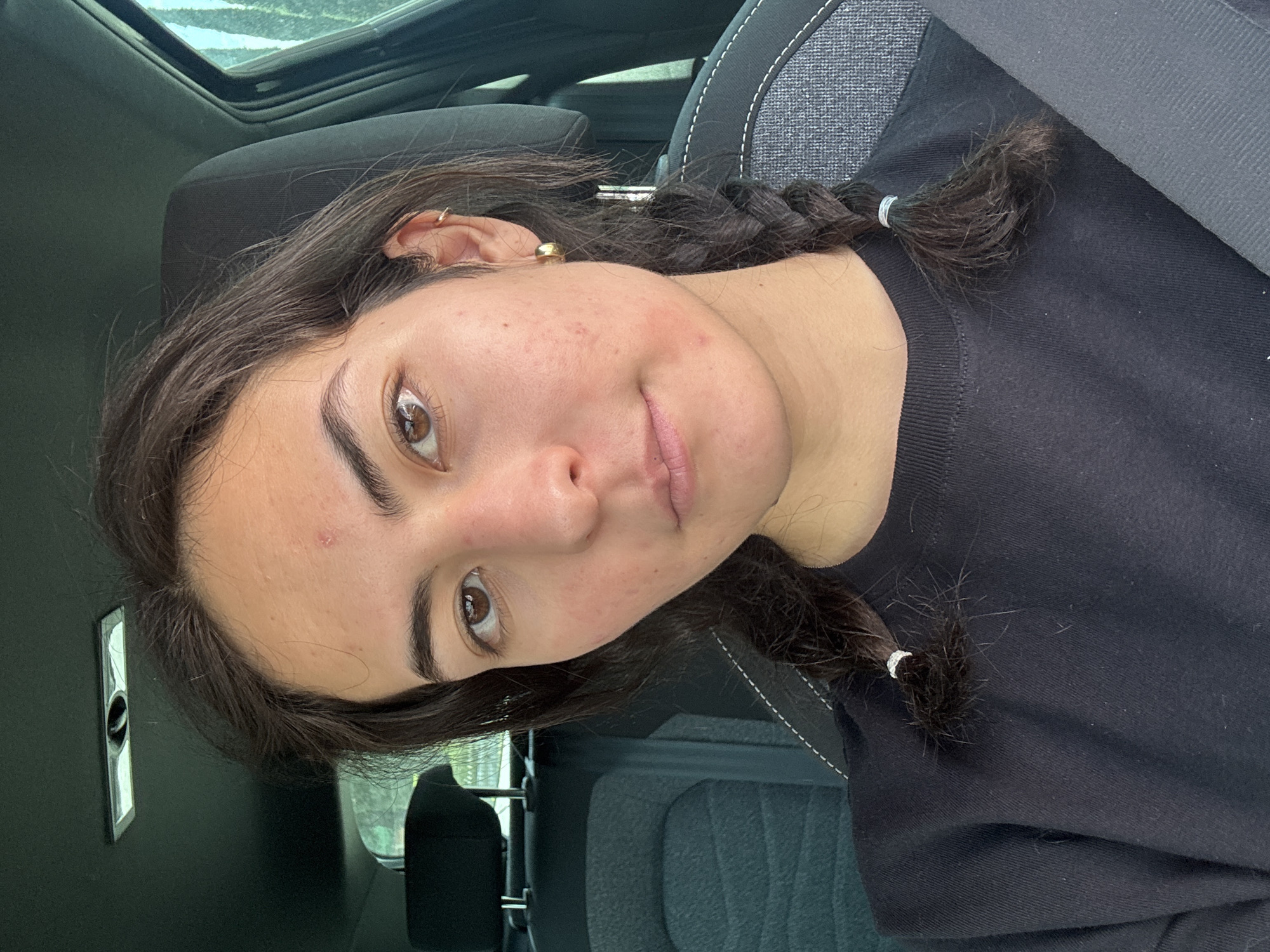 A young woman with brown hair braided into two pigtails is shown from the chest up, slightly tilted, within a car interior. She is wearing a black t-shirt and a seatbelt. The car has a sunroof and grey upholstery. The location in the city of San Bernardino Tlaxcalancingo, Mexico is not determinable from the image.