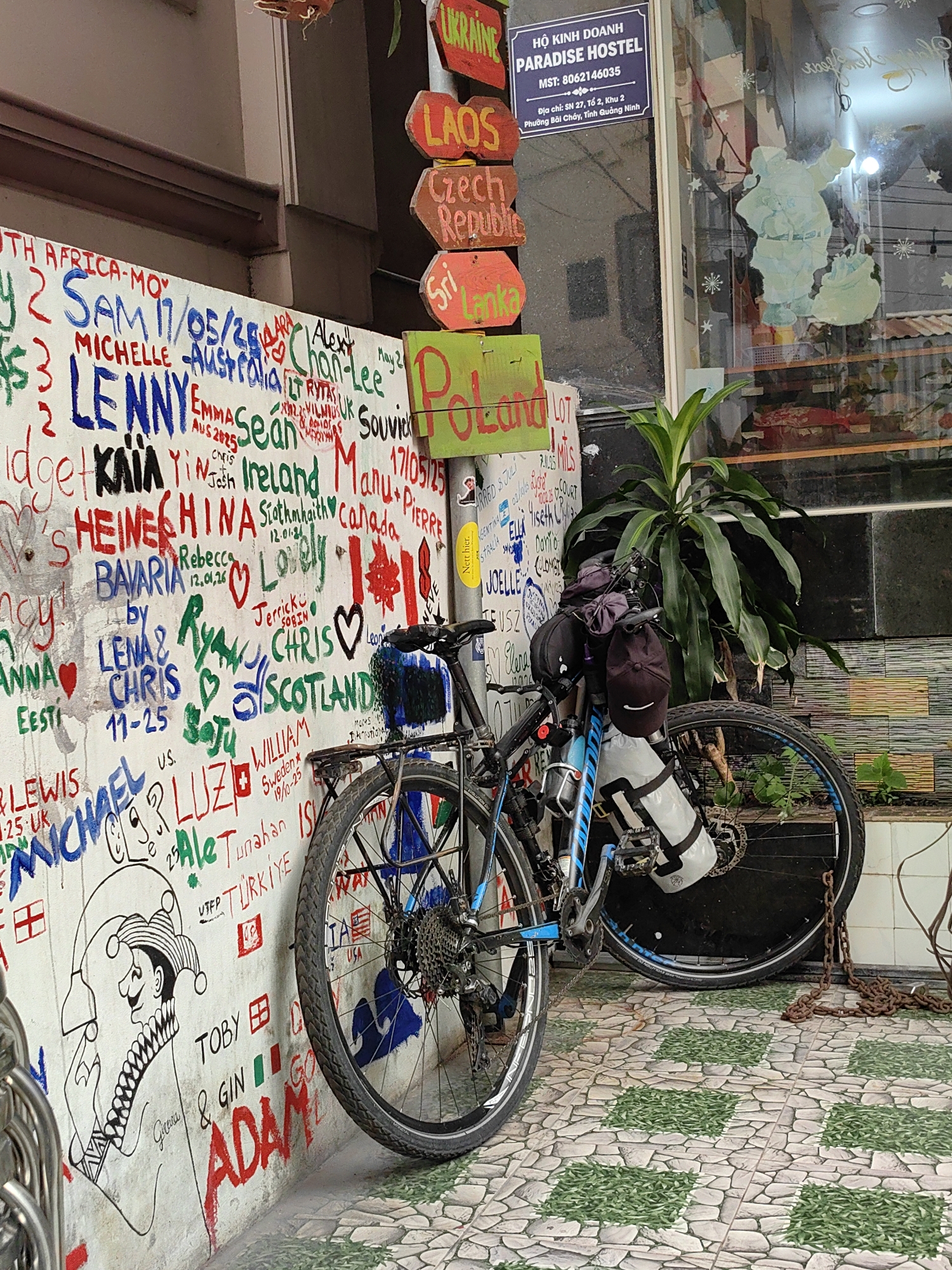 The image depicts a bicycle parked against a wall covered in graffiti. The wall includes writings in various colors and languages, listing names, dates, and countries like "Ukraine," "Laos," "Czech Republic," "Sri Lanka," "Poland," "China," and "Canada." A drawing of a jester is also visible.
Next to the graffiti wall is a sign indicating "Paradise Hostel" with the address being SN 27, Tổ 2, Khu 2, Phường Bãi Cháy, Tỉnh Quảng Ninh, suggesting the hostel is located in Bai Chay Ward, Vietnam. The bicycle is a touring model with loaded panniers and water bottles. There is a plant near the bicycle, and the floor is tiled with a pattern resembling stone with leafy insets. A chain is on the ground nearby. A window to the hostel is visible in the background, partially obscured by decorations.