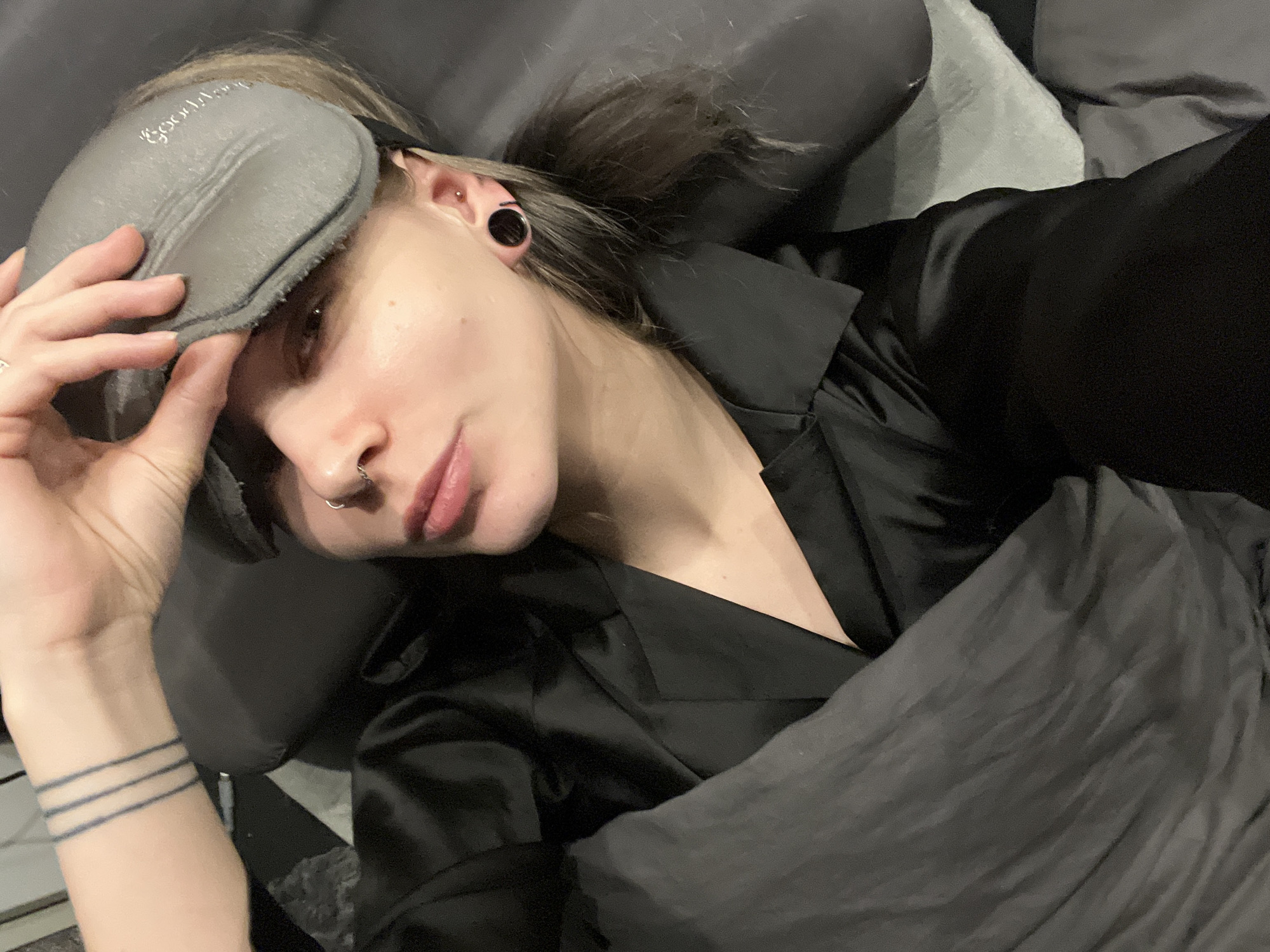 A person is shown in a medium close-up shot, lying in bed and looking directly at the camera. They have light-toned skin, dark hair pulled back, and dark eyes. A grey sleep mask, with the word "GoodNight" in white script, is pushed up onto their forehead and held by their left hand.
Notable details include several piercings: a silver septum ring and a small ring piercing on the side of their left nostril. Their left ear features a black ear tunnel (gauge) and a small silver stud piercing higher up. On the person's left wrist, three thin, parallel dark lines are tattooed. They are wearing a black collared shirt made of a satin-like material, possibly pajamas. The person is partially covered by dark grey bedding and resting on a dark grey pillow. The scene takes place indoors, likely in a bedroom, and the activity suggests resting or waking up. The expression is neutral and direct.