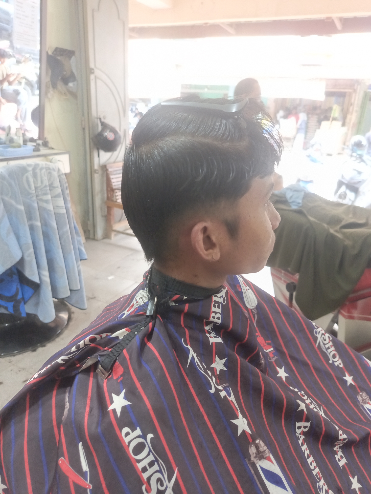 The image captures a young male, likely a teenager, from the shoulders up, in a barbershop in Lhokseumawe, Indonesia. He is wearing a striped barber cape with stars and razor patterns, predominantly in dark blue, red, and white. His hair is styled with a comb on top, indicating he is in the process of receiving a haircut. The background reveals a bright, airy interior with some outdoor elements visible through a doorway or window. To the left, a table with a light-colored cloth and a blue basin is present. Further back, wooden chairs and a rack with helmets suggest a busy environment. The overall lighting is bright, with some overexposure in the upper portion of the image, likely from natural light.