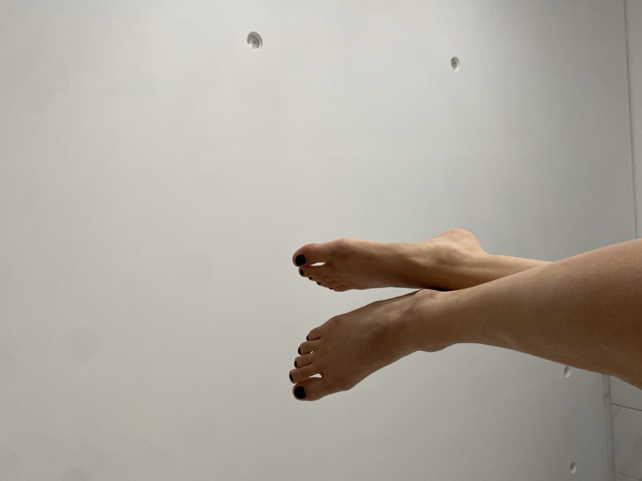 A pair of human legs and bare feet are depicted, extended upwards and crossed at the ankles, positioned against a plain white ceiling. The toenails on all visible toes are painted black. Two circular recessed light fixtures are visible in the upper right quadrant of the ceiling. A faint light-colored surface is partially visible at the bottom left corner of the frame. The scene is located indoors in Skala, Greece.