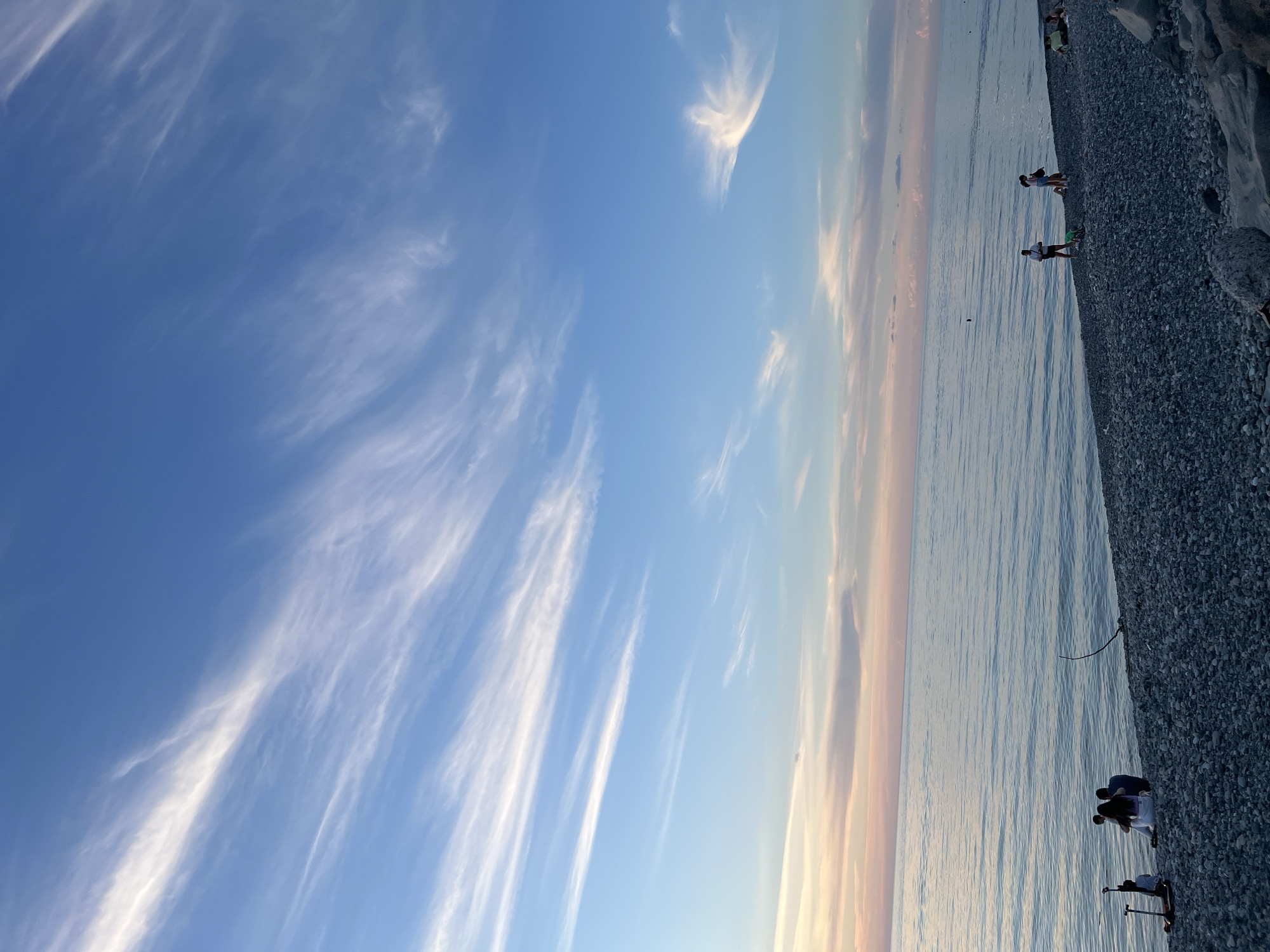 This image depicts a serene beach scene at sunset in Batumi, Georgia. The sky is a vibrant canvas of blue with streaks of wispy white clouds, transitioning to soft hues of pink and orange near the horizon, indicating the sun is setting over the Black Sea. The sea itself is calm, with gentle ripples reflecting the sky's colors.
The beach is covered in smooth, grey pebbles. In the foreground, on the left side of the frame, a group of people is gathered, some sitting and others standing, with a scooter nearby. Further along the beach, towards the right, several other individuals are seen, some walking and others sitting. A small dog on a leash stands with one of the beachgoers. The overall atmosphere is one of relaxation and enjoyment of the twilight.
