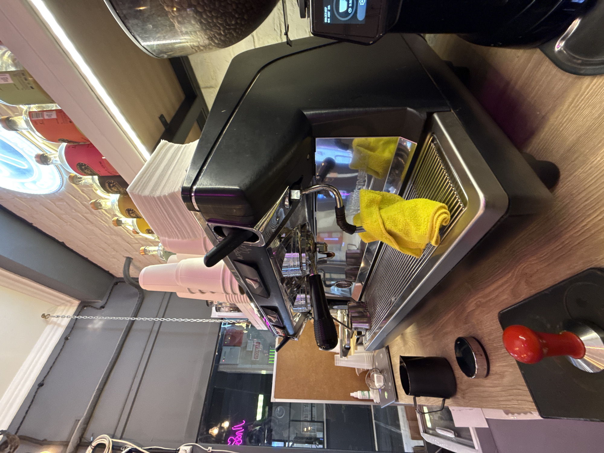 In an indoor commercial setting, an espresso machine, likely in a cafe or coffee shop, is the central subject. The machine is primarily black and silver, with a prominent portafilter, steam wand, and drip tray. A yellow microfiber cloth is draped over the drip tray, and a metal milk pitcher sits to the left of the machine. On the counter in the foreground, a red-handled tamper rests on a black mat. A black pitcher and a silver oval object are also on the counter.
Behind the espresso machine, a shelf displays several bottles, some with colorful labels, and stacks of white paper cups and napkins. A large glass container filled with coffee beans is visible to the right of the espresso machine, part of a coffee grinder. The background reveals a brick wall and a window with reflections of lights and possibly other customers or elements of the cafe interior, including a pink neon sign. The scene appears to be a well-lit, functional workspace. The location is identified as Unknown, Unknown.