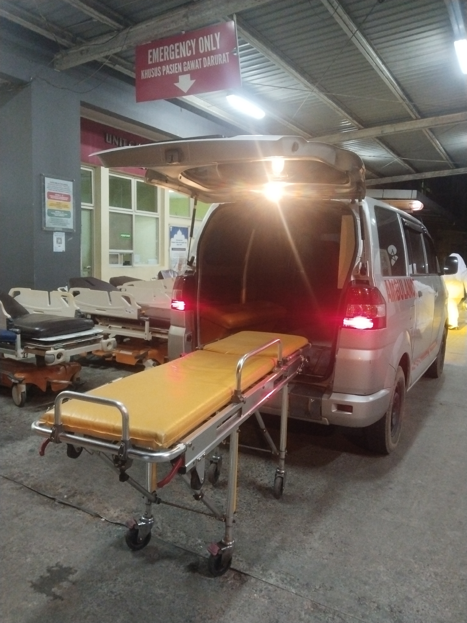 The image shows an ambulance parked near a building in Banda Aceh, Indonesia. The ambulance's rear door is open, revealing a yellow-colored stretcher partially extending out. A red sign overhead reads "EMERGENCY ONLY KHUSUS PASIEN GAWAT DARURAT" with a downward pointing arrow. Additional stretchers are visible near the building's entrance.
