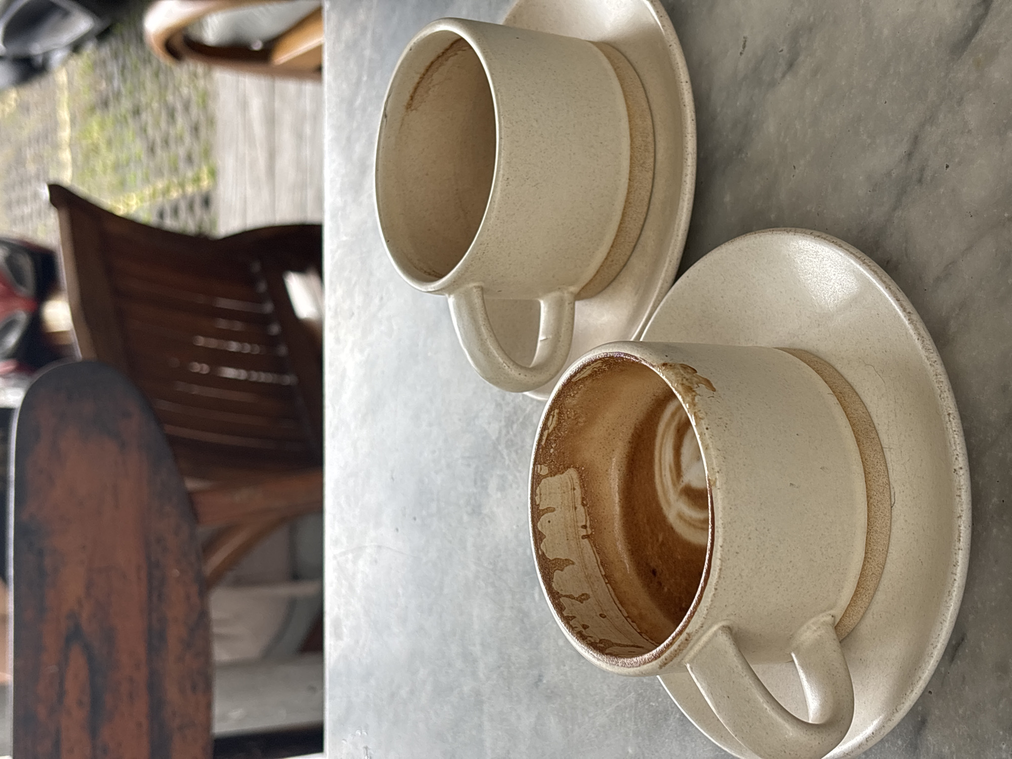 The image captures two empty ceramic coffee cups with their accompanying saucers, resting on a mottled gray surface, likely a stone or marble table, in an outdoor setting in Kuta Selatan, Indonesia. The cups are a light, speckled beige. One cup is inverted on its saucer, displaying its exterior and handle. The second cup is positioned upright but tilted, revealing dried, reddish-brown residue of a consumed beverage, presumably coffee or tea, adhering to its inner walls and forming a circular pattern at the base. Both cups feature a curved handle.
The background is softly blurred, showing elements of an outdoor area. A dark wooden chair with a slatted backrest is visible, along with hints of other wooden furniture, greenery, and a patterned ground surface. The lighting suggests it is daytime. There are no people visible, and the scene depicts the quiet aftermath of a meal or break, with the empty cups indicating that beverages have been recently consumed. No text is visible within the image.