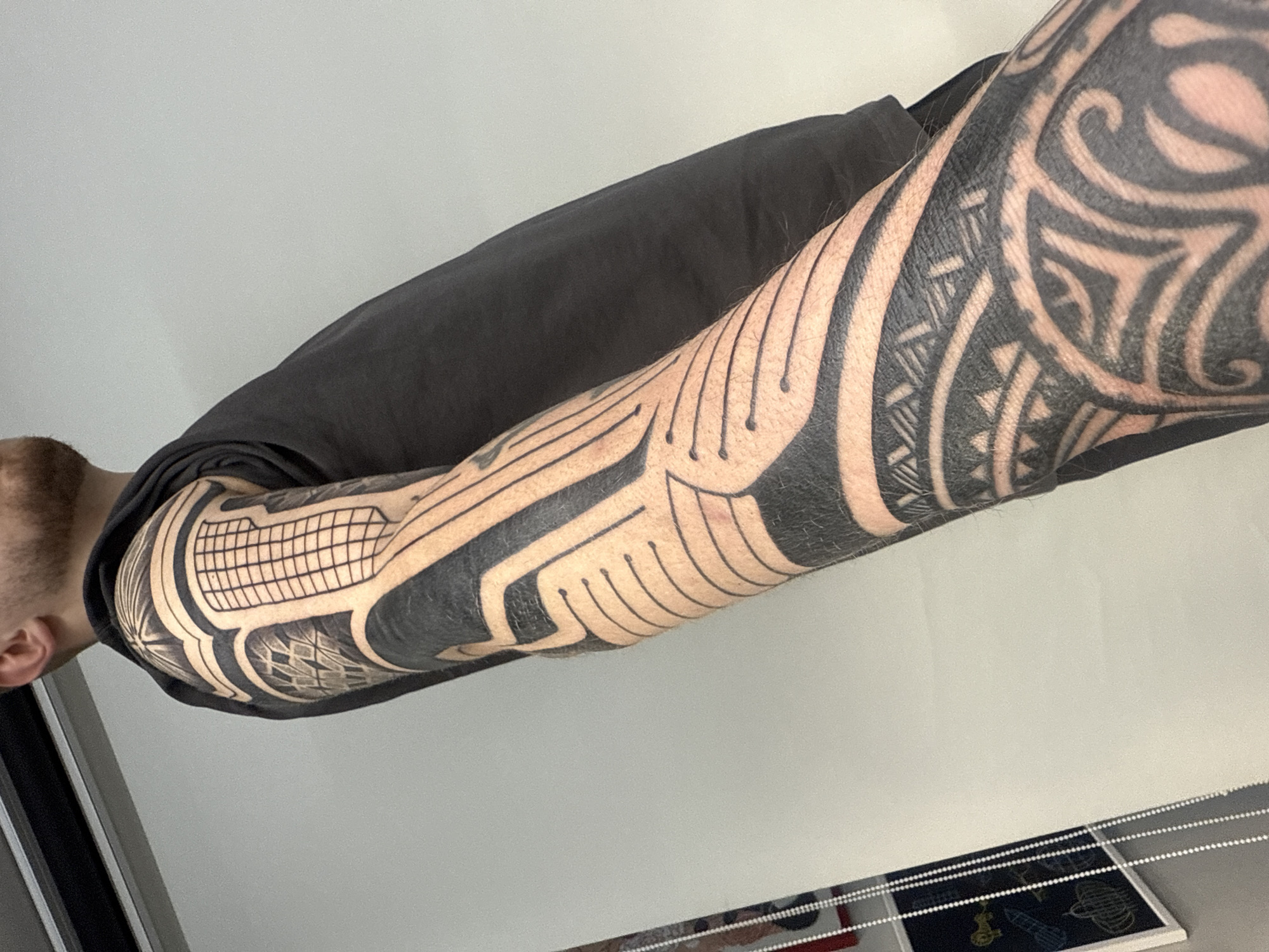 A man wearing a black t-shirt is seen with his heavily tattooed left arm extended. The tattoo designs are elaborate, featuring thick black tribal-inspired patterns and geometric shapes. The setting appears to be indoors, with a white wall in the background. The specific location within Sofia, Bulgaria cannot be determined from the image alone.