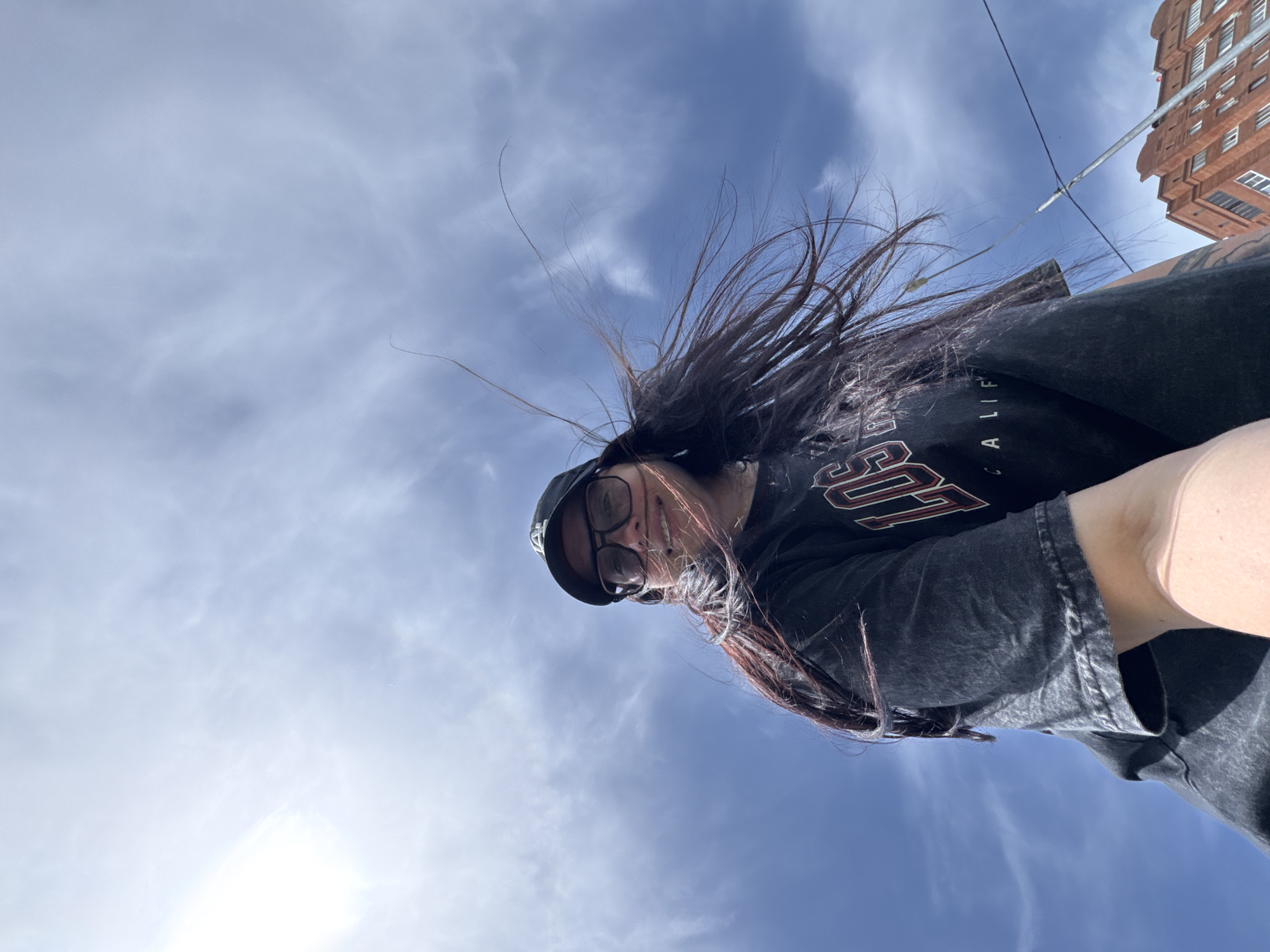 A low-angle shot shows a person in a black baseball cap and glasses, their dark hair blowing upwards against a cloudy blue sky. The person is wearing a black t-shirt with white and red lettering. Part of a brick building can be seen in the upper right corner. The location is determined to be in an unknown city of unknown.