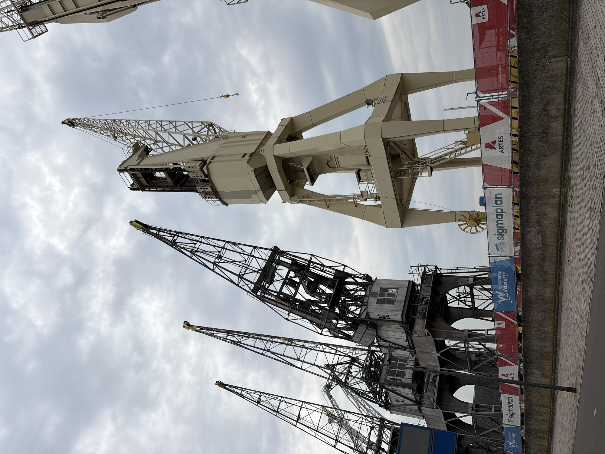 The image shows a view of cranes in the Port of Antwerp, Belgium. The cranes are of varying design; some are older, with a dark, lattice-like structure, while others are newer and painted beige. The cranes are located near a concrete wall, with signage bearing the name "ARTES" and "sigmaplan." The sky above is overcast with grey clouds. The image is oriented vertically.