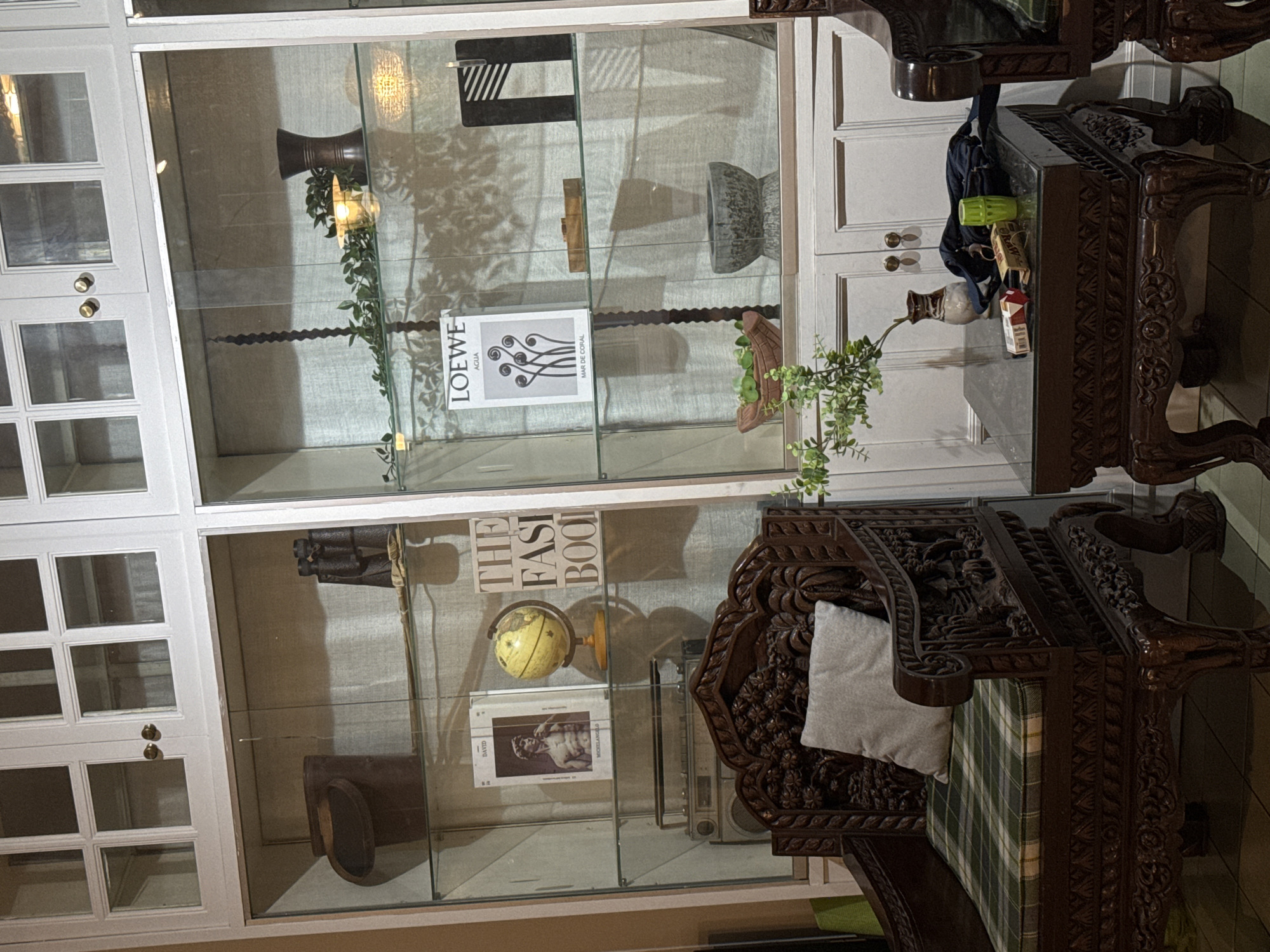 The image shows a display cabinet filled with various objects, including books, a globe, a vase, and decorative items. The cabinet is made of white wood with glass doors and shelves. On the left side of the cabinet, there is a brown bucket, a pair of binoculars, and a book about Michelangelo's David. In the center, there is a yellow globe and a book titled "The Fasi Book." On the right side, there is a LOEWE advertisement, a wooden sculpture, a black and white artwork, and a bowl. Below the cabinet, there is a vintage boombox and a carved wooden chair with a green and white plaid cushion and a light gray pillow. A small dark wood table with a glass top is placed in front of the chair. On the table, there is a vase with a small plant, a pack of cigarettes, a lighter, a green cup, and a black bag. The setting appears to be an indoor living space in Chiang Mai City Municipality, Thailand, possibly a home or a shop. The lighting is dim, with spotlights illuminating the items in the display cabinet.