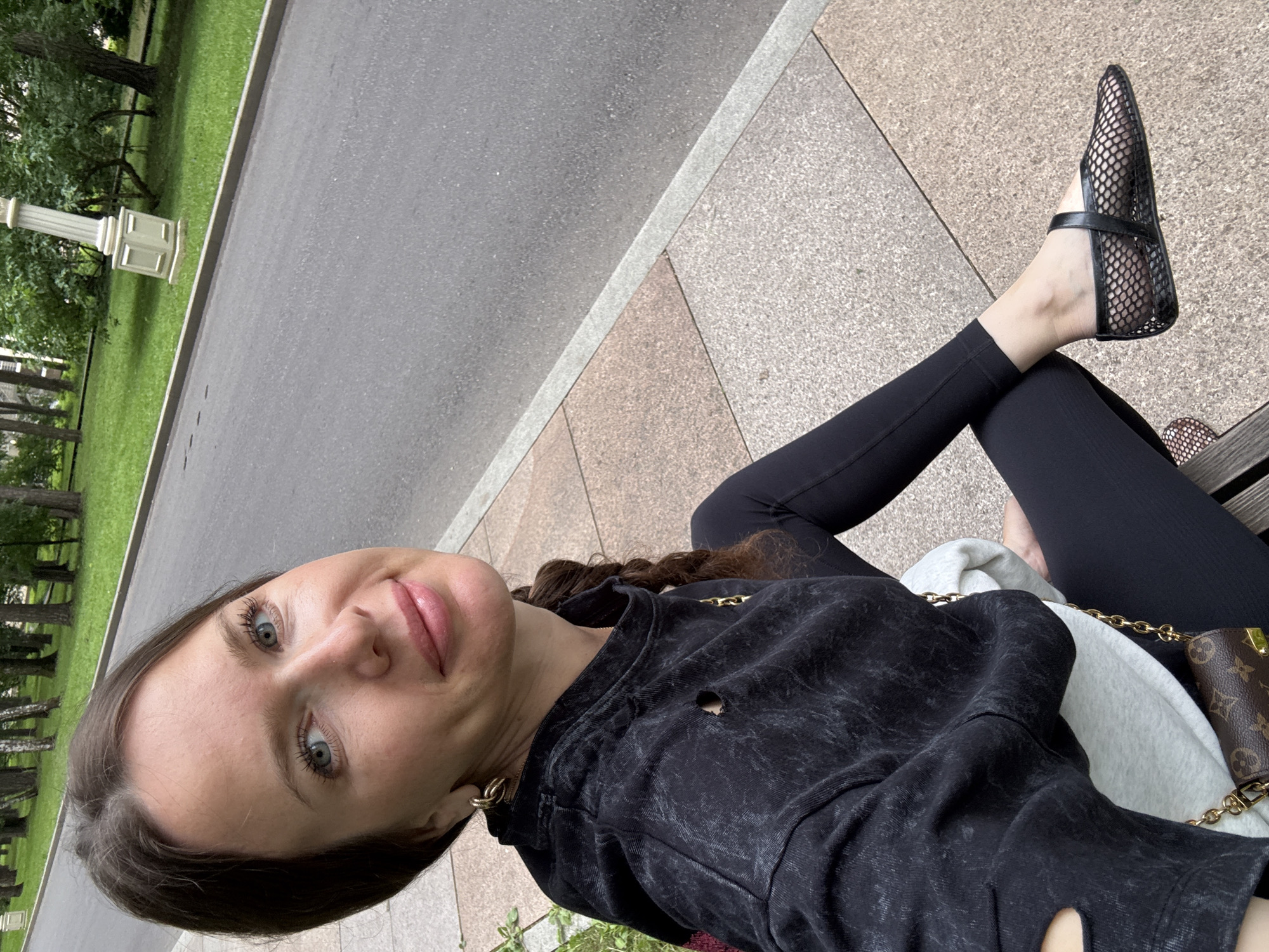 The image shows a young woman sitting on a bench outdoors, with a path and grassy area behind her. The woman is wearing black leggings, a dark grey, acid-washed sweatshirt with a hole on the left side, and black mesh ballet flats with a strap. She has long brown hair, blue eyes, and is wearing gold hoop earrings. A small brown designer handbag with a gold chain is visible on the bench next to her. The overall lighting suggests it is daytime, with overcast or diffused sunlight. The presence of trees and a manicured grassy area, along with a paved path and what appears to be a lamppost, indicates a park or outdoor recreational space. The provided location context is Moscow, Russia. The woman is looking towards the camera, with a slight smile, conveying a relaxed and casual mood. No visible text is present in the image.