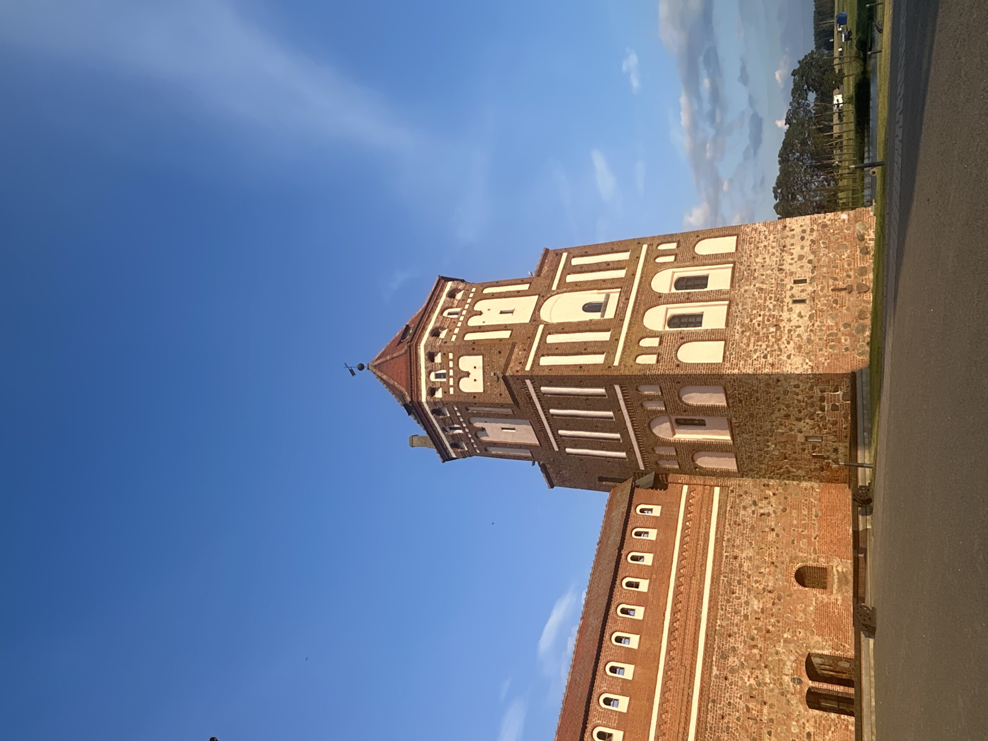 The image depicts a tall, octagonal brick tower with a red tiled roof, part of a larger red brick castle complex. The tower has a weathered stone base and is decorated with white arched windows and vertical white rectangular accents. A long, low wing of the castle, also constructed of red brick with a red tiled roof, extends to the left, featuring a series of evenly spaced white arched windows. The castle is set against a bright blue sky with wispy white clouds and some distant trees. In the foreground, there is a paved area, likely a courtyard or pathway, and the edge of a grassy area with some distant vehicles. The lighting suggests it is either early morning or late afternoon, with long shadows cast on the ground. This scene is located at the Mir Castle Complex in the village of Mir, within the Zhukhavitski Sel'ski Savet of Belarus.