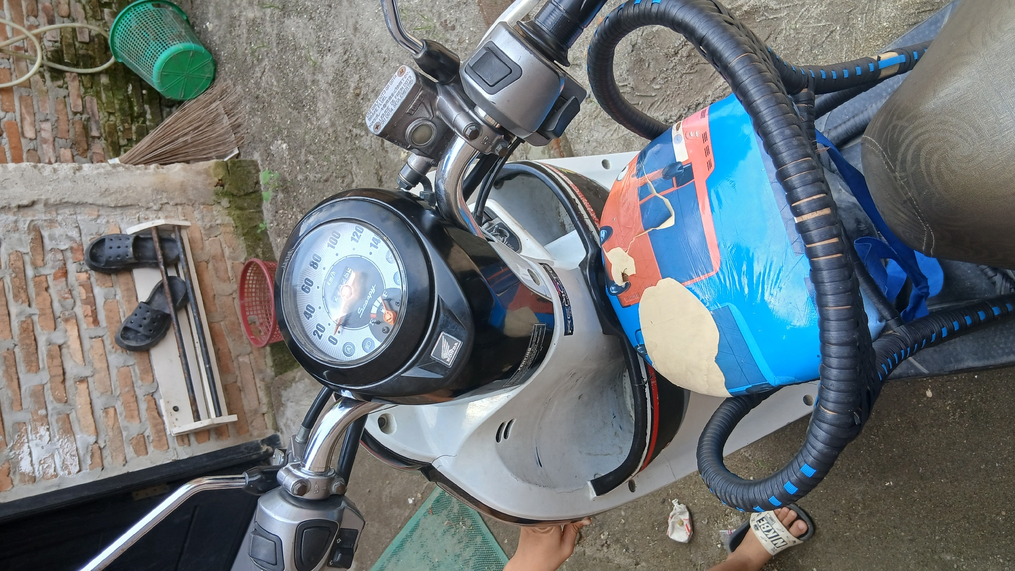 The image shows a close-up view of a white and black Honda Scoopy motorcycle parked outdoors in what appears to be a residential area. The motorcycle is the primary subject, with its handlebars, instrument cluster, and front bodywork clearly visible. The instrument cluster shows a speedometer with a needle indicating a speed below 20 km/h. A person's left hand is seen reaching towards the motorcycle's bodywork. A small child's left foot wearing a white sandal with "NIKE" written on it is visible in the lower left corner. In the background, a brick wall with a white shelf holding two black flip-flops is present. To the right of the wall, a broom and a green plastic trash bin are also visible. The ground is a mixture of concrete and loose soil. The scene is consistent with an outdoor setting in a moderately maintained urban or suburban environment, possibly a yard or alleyway in Klambir Lima Kebun, Indonesia, given the overall aesthetic and objects present.