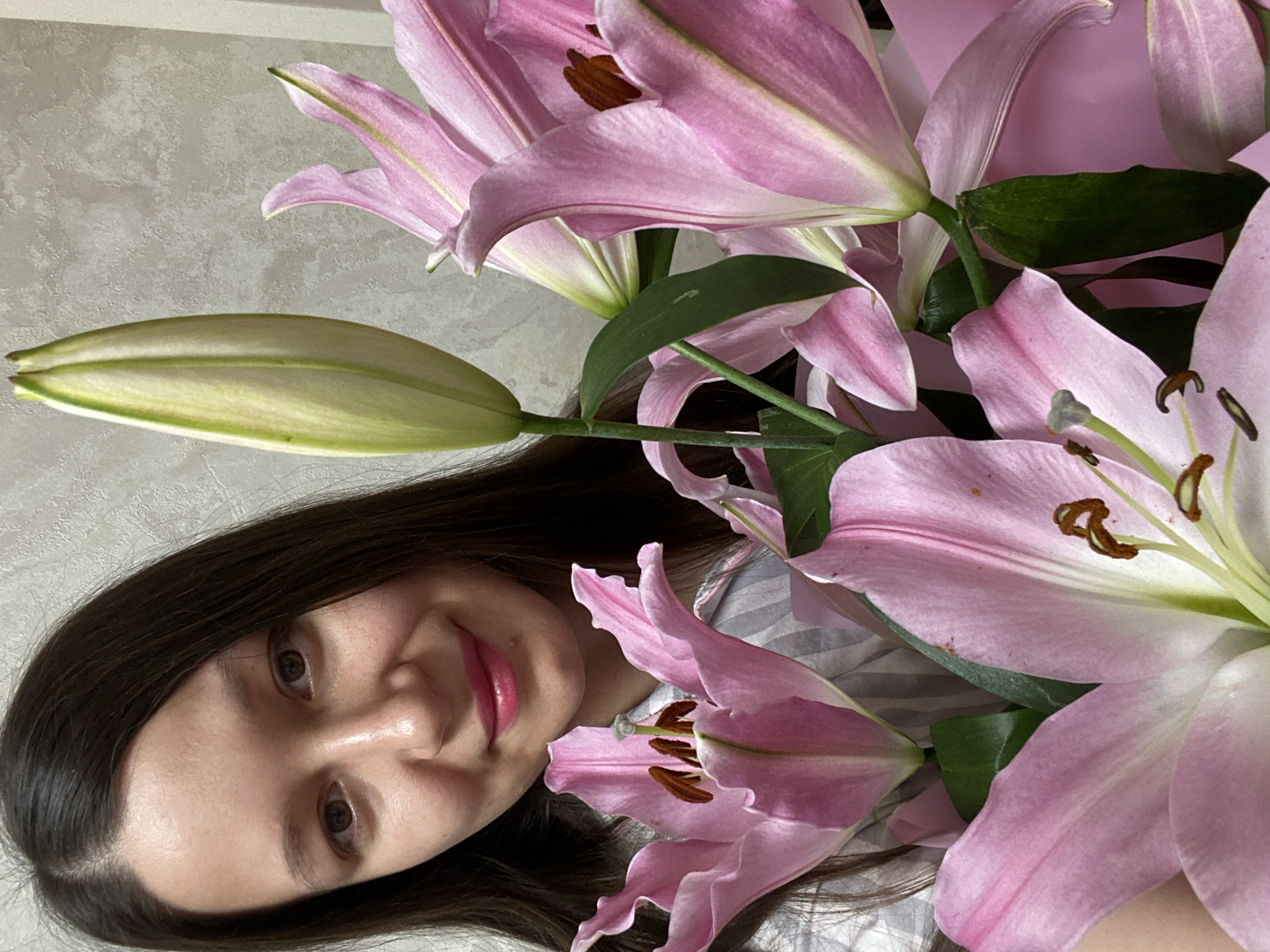 A young woman with dark hair and light skin smiles into the camera from slightly below, surrounded by a bouquet of pink lilies. The lilies feature prominent stamens with brown tips, and some are still in bud. The woman is wearing a striped top. A wall with a textured, gray surface is visible in the background. The location is Chaikovsky, Russia.