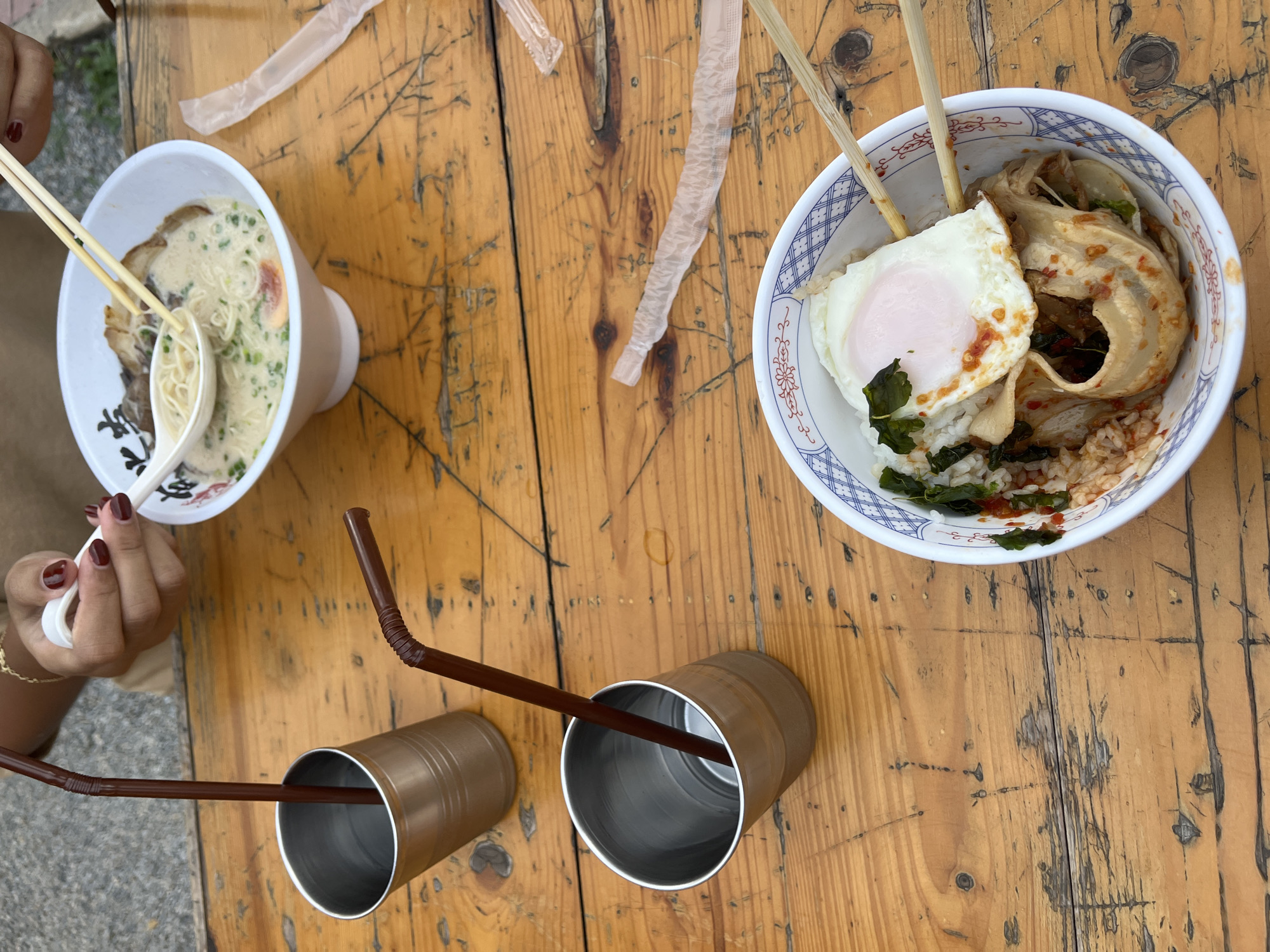 The image depicts a close-up, overhead view of a wooden table setting at an outdoor restaurant in Chiang Mai City Municipality, Thailand. Two bowls of food and two metallic cups with brown straws are arranged on the rustic, weathered wooden surface. In the foreground, a bowl contains rice topped with a fried egg, some greens, and a meat dish, being held by chopsticks. To the left, a metallic cup with a straw is positioned near the edge of the frame. In the background, another bowl, filled with ramen, is visible, with chopsticks and a spoon in use. A person's hands are visible in the upper right, holding a spoon and chopsticks. The overall scene suggests a casual meal being enjoyed outdoors.