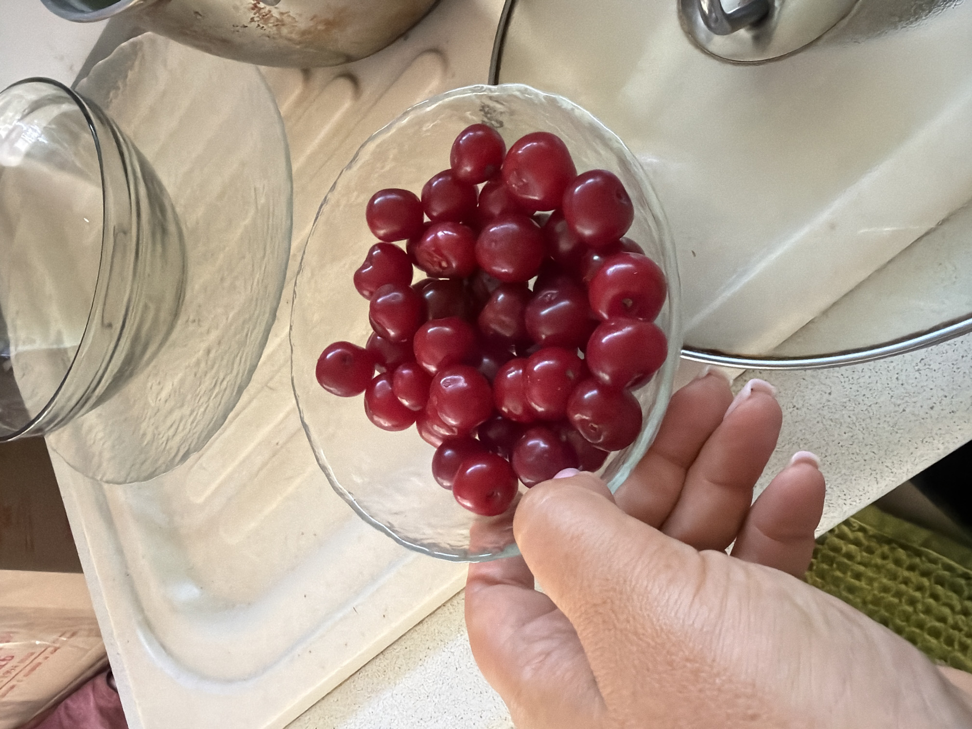 A close-up shot shows a hand holding a clear glass bowl filled with ripe cherries. The cherries are a deep red color and appear fresh. The bowl is partially full, with many cherries visible. The hand is positioned on the left side of the frame, with fingers slightly curled around the bowl. The background consists of a kitchen counter with a light-colored surface. A hint of a green textured item is visible at the bottom left of the image. The scene is indoors, likely in a home kitchen in Leningradskaya, Russia.