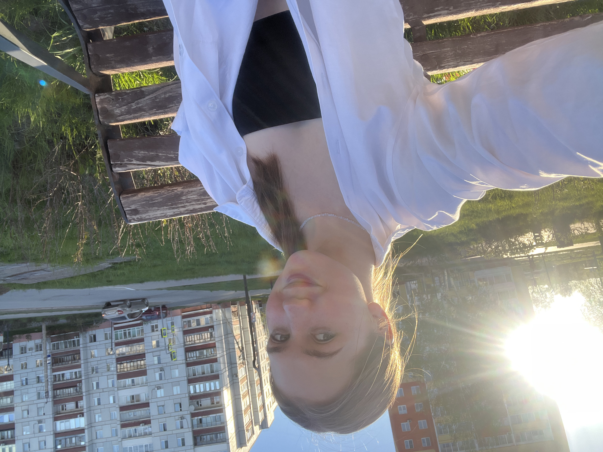 A young woman is taking a selfie. She is wearing a white, button-down shirt over a black top. She is sitting on a wooden bench. Behind her are apartment buildings, indicating an urban setting. Cars are visible on a road near the buildings. The sun is brightly shining, positioned behind some trees and buildings. Based on the architectural style of the buildings, this image appears to be taken in a Russian city, likely Chaikovsky as specified.