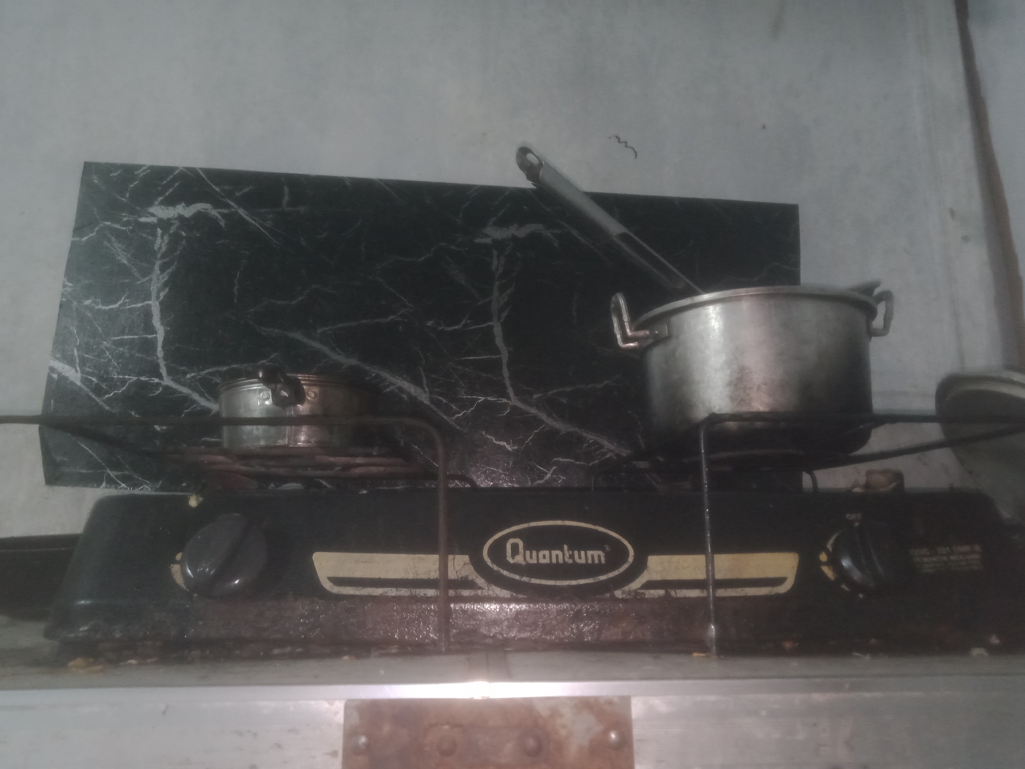 The image shows a two-burner gas stove with pots on top. The stove is black with a marble-patterned backsplash behind it. The brand name "Quantum" is visible on the stove. The stove is located on a metal shelf. The background appears to be a plain wall. The lighting is dim, suggesting an indoor setting. The stove appears to be in a kitchen or cooking area.