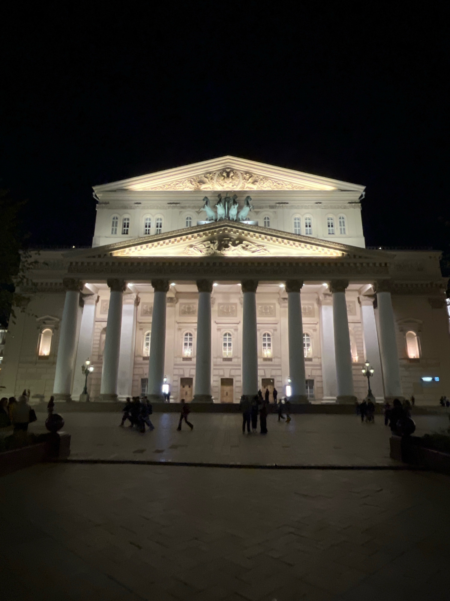 The image shows the Bolshoi Theatre in Moscow, Russia at night. The grand facade of the theatre is illuminated, highlighting its neoclassical architecture with large columns and a pediment adorned with sculptures. Several people are visible on the plaza in front of the theatre, some walking individually and others in small groups. The plaza is paved and appears to be a public space. The sky is dark, indicating it is nighttime.