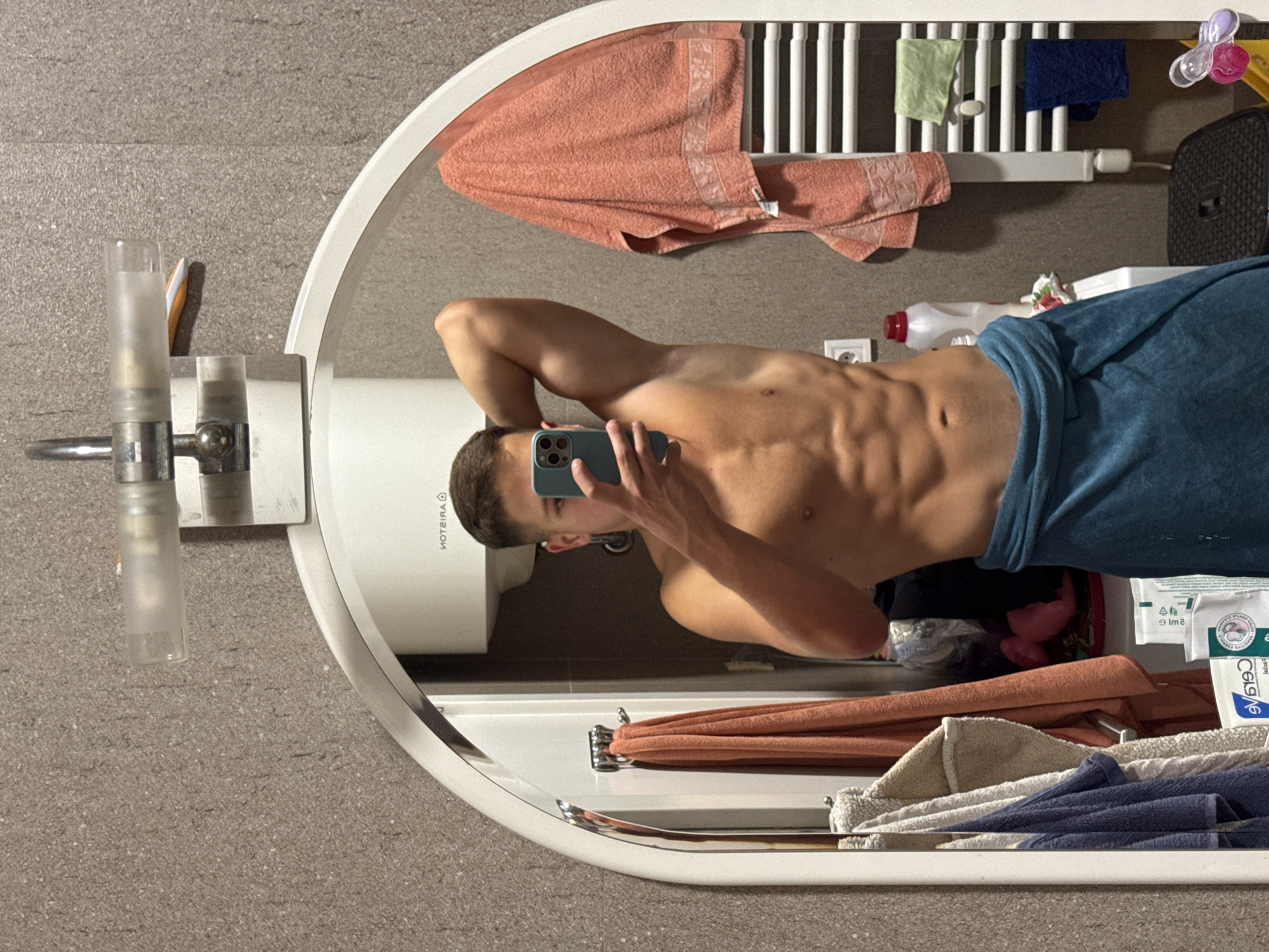 A young man with a towel wrapped around his waist poses for a mirror selfie in a bathroom. He is shirtless, revealing well-defined abdominal muscles and arm muscles. He holds a smartphone in his right hand, angled to capture his reflection. The bathroom environment includes a wall-mounted heater with several towels draped over it, various toiletries, and a sink. The overall aesthetic suggests a domestic setting, possibly a hotel room or personal residence, and the image does not contain any elements that would indicate a specific location within the city of Budva, Montenegro.