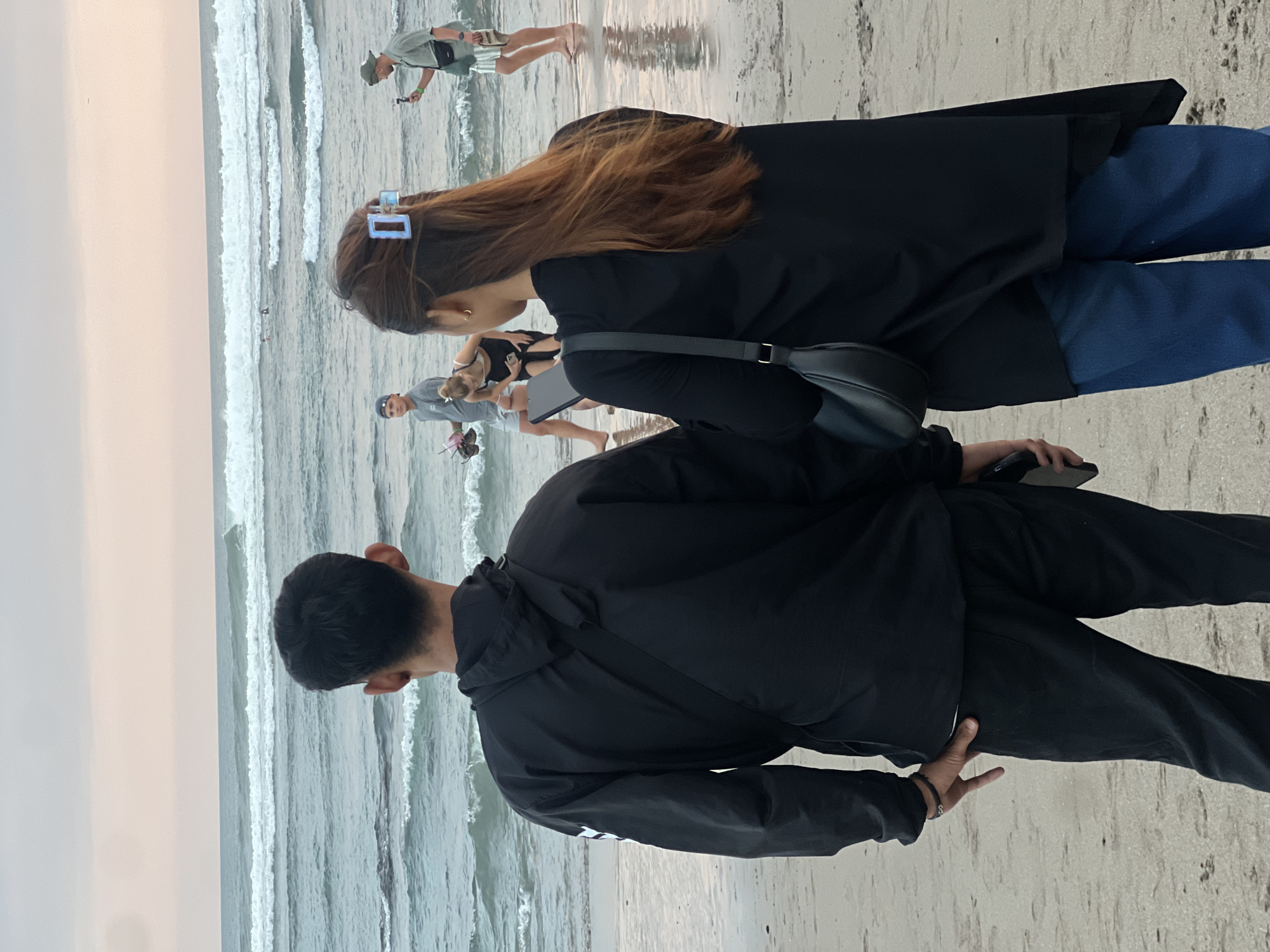Two people, a man and a woman, stand on a sandy beach facing the ocean in North Kuta, Indonesia. The man, from the back, wears a black hooded jacket and black pants. A dark strap from a bag is visible across his back. The woman, also viewed from behind, has long brown hair secured with a blue clip. She wears a black top and blue jeans. She holds a black phone in her right hand. Behind them, several other individuals are present near the water's edge. One man, wearing a light-colored shirt and shorts, holds a phone and appears to be filming. Another woman in a white top and dark shorts is bending over. Further out in the ocean, waves are breaking, and a few people are visible in the water, possibly surfers. The sky is overcast with soft hues of pink and orange, suggesting it might be late afternoon or early evening. The sand is damp and reflects the light near the water.