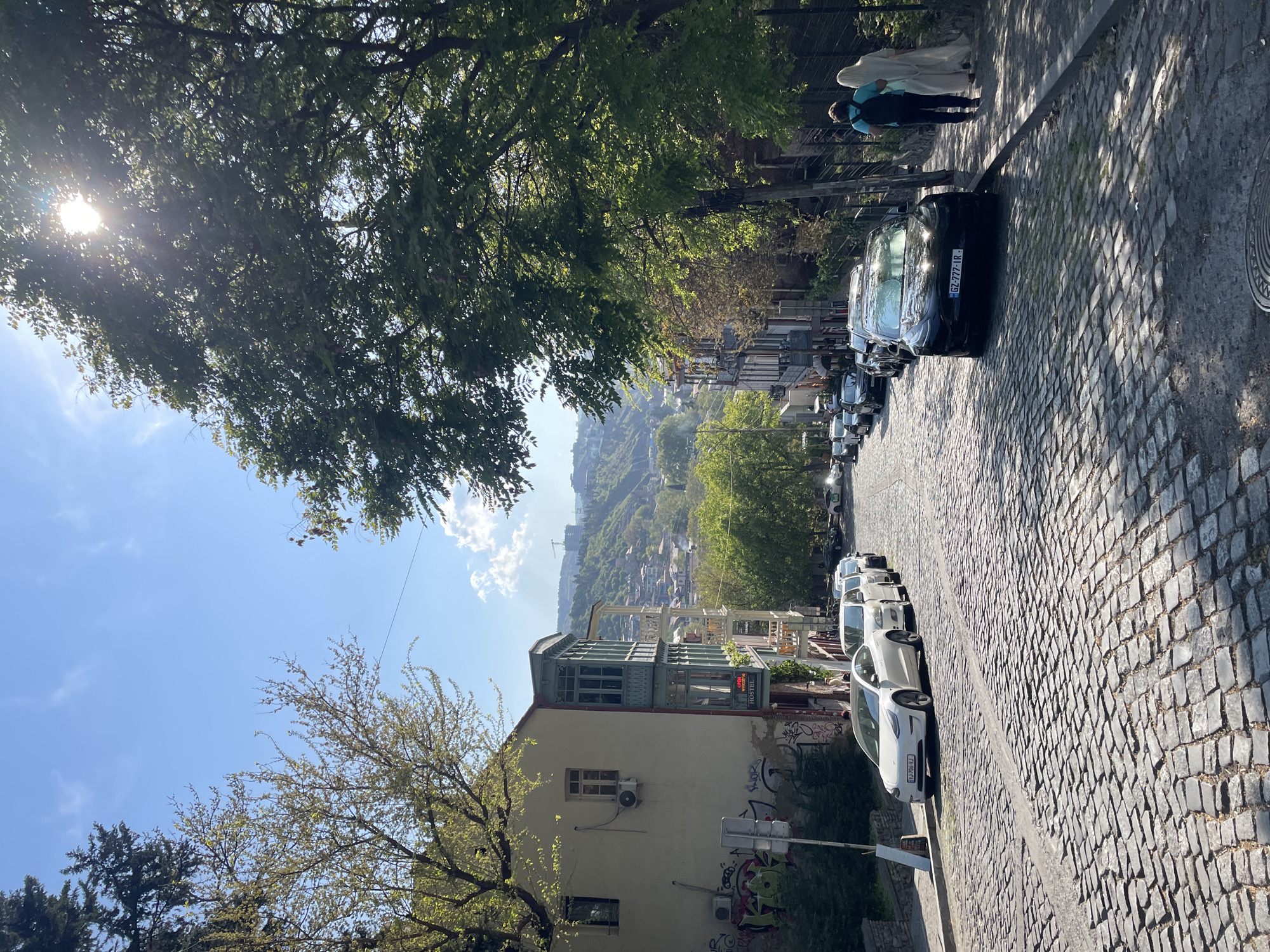 The image portrays a sun-drenched, descending cobbled street in an urban hillside environment, consistent with Tbilisi, Georgia. On the right side of the street, several cars are parked; prominent among them is a black car with the license plate "GZ-777-IR" and further down, white cars, one displaying "IO-796-XJ". Two people are walking on the sidewalk at the upper right; one wears a light blue top and dark pants with a backpack, while the other is clad in a long, light-colored garment and a head covering.
To the left, a building stands out with its distinctive blue-green enclosed wooden balcony or veranda. The building's lower wall is light-colored and adorned with graffiti, and an air conditioning unit is visible. A sign on this building clearly reads "HUEB WINDOW HOSTEL COFFEE". The street is canopied by abundant green trees, allowing bright sunlight to dapple through the leaves and cast long shadows across the cobblestones, indicating midday or early afternoon.
In the background, a panoramic view of a sprawling cityscape stretches across undulating hills, densely packed with buildings and hints of distant construction under a clear blue sky with scattered white clouds. The scene conveys a calm, everyday atmosphere.