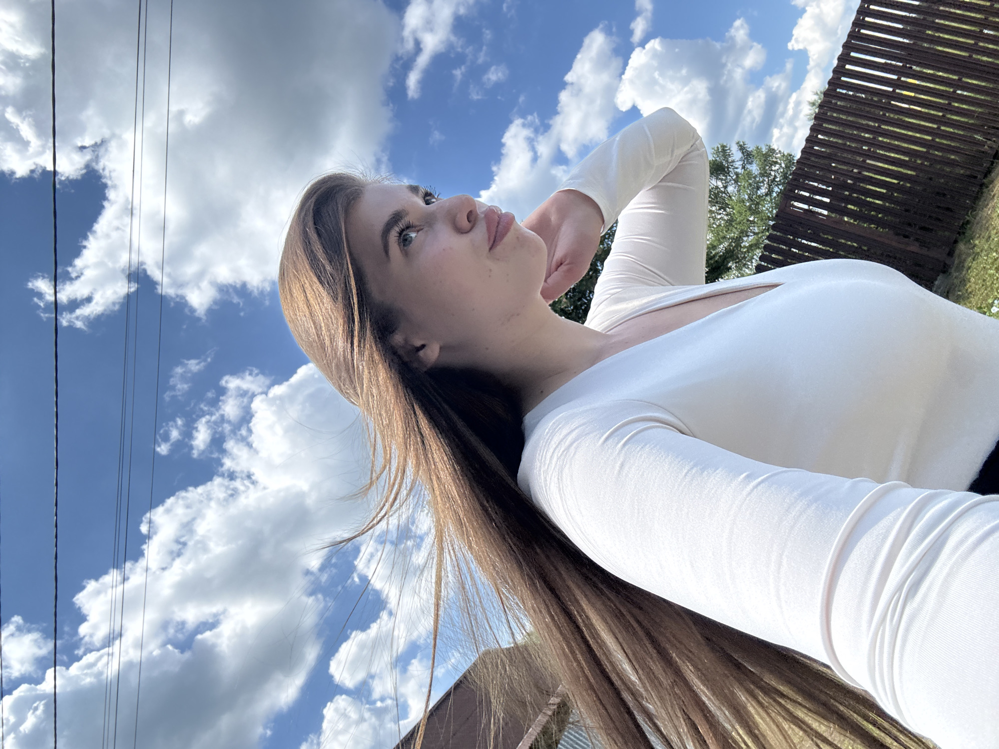 A young woman with long, brown hair stands outdoors, her hair blowing in the wind against a backdrop of a bright blue sky with white, fluffy clouds. She is wearing a white, long-sleeved shirt with a V-neckline. She is looking off to her right, her right hand touching her chin. In the background, a wooden fence and some greenery are visible. The scene appears to be in a rural or suburban area. There is no indication that the image was taken in the city of Мещёры, Russia.