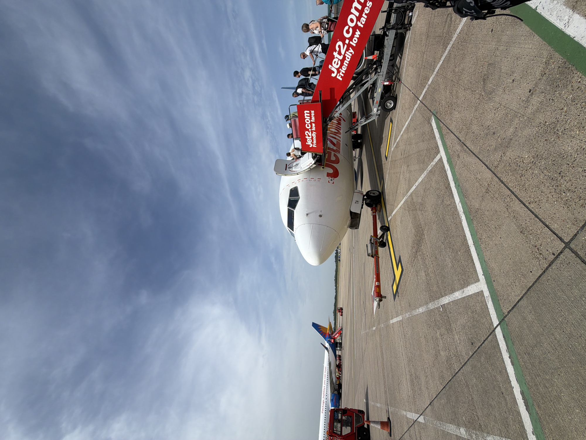 The image depicts a Jet2.com passenger airplane at an airport, likely in Uttlesford, United Kingdom. Passengers are disembarking using a set of mobile stairs bearing the Jet2.com logo. The aircraft has a white fuselage with red and yellow Jet2.com branding. The sky is partly cloudy. Several ground vehicles are visible on the tarmac. Another airplane, partly visible in the distance, has a tail fin with a blue and yellow design. The ground is concrete with painted white and green lines delineating parking and movement areas.