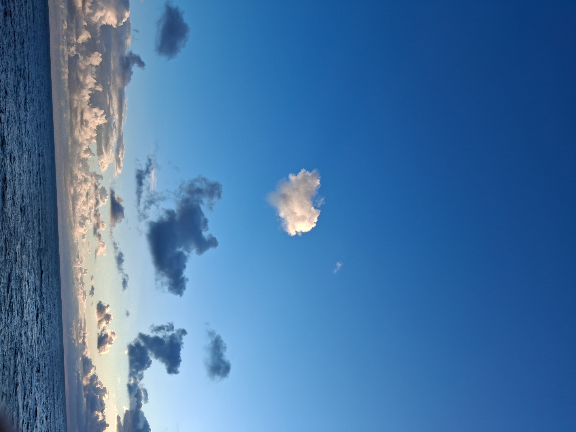 The image shows a view of the sky and the ocean, presumably taken in Figueira da Foz, Portugal. The horizon is to the left of the frame, showing the ocean. A line of clouds is visible along the horizon, with colors transitioning from light to dark. The sky above the clouds is predominantly blue, with some scattered dark and white clouds.